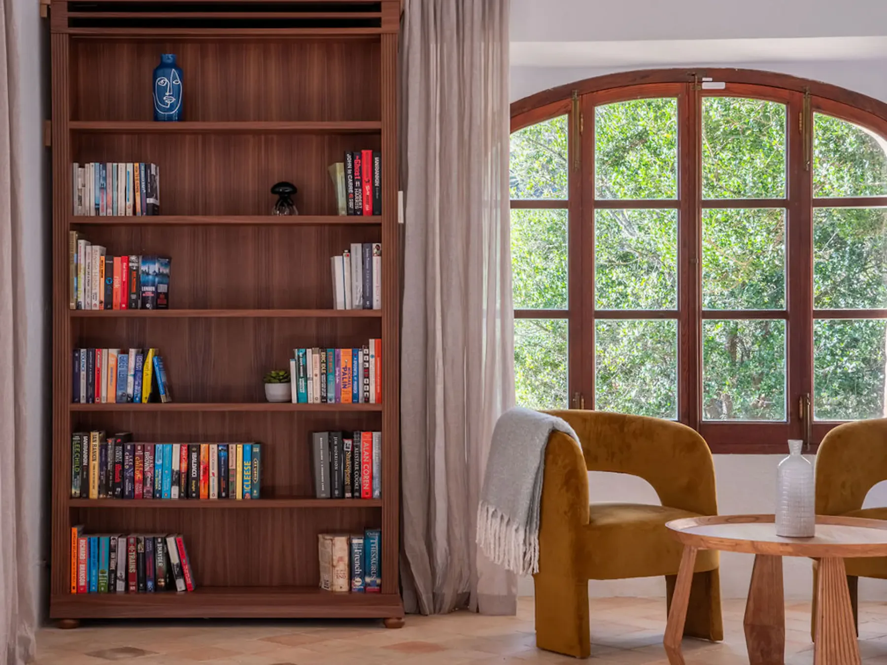 A reading nook with a wooden bookcase beside an arched window and seating.