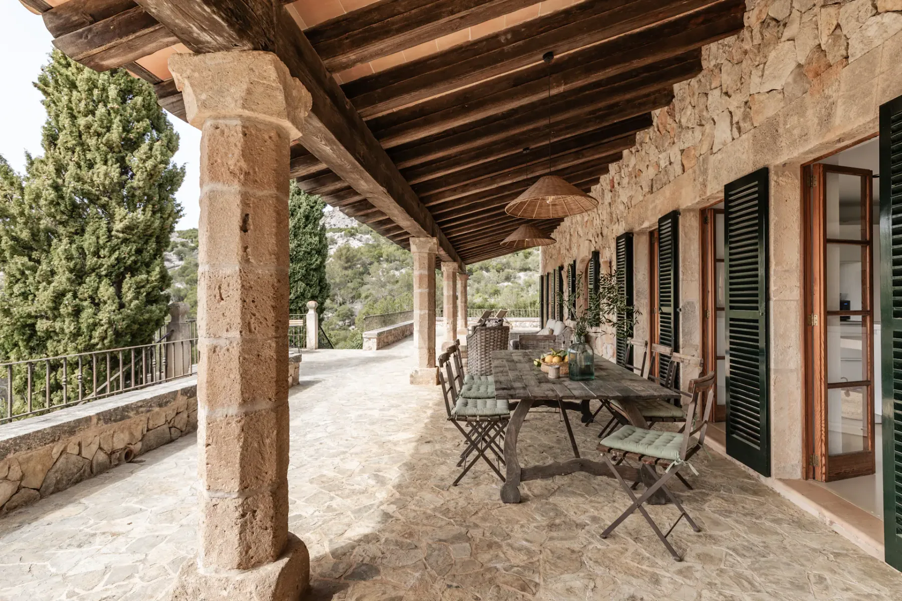 A covered stone terrace has a long table and seating beside shuttered windows.