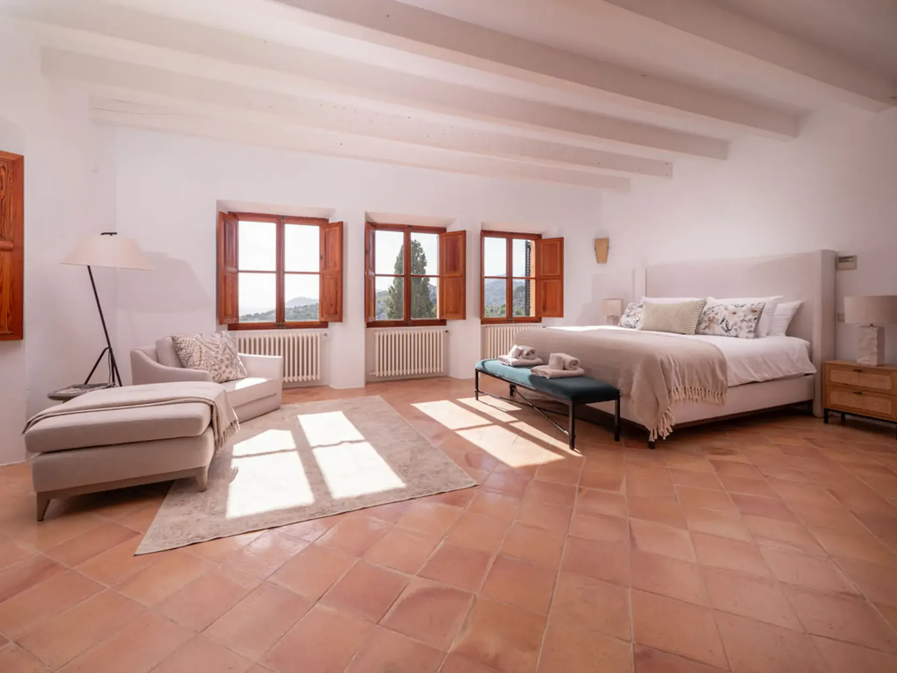 A bright bedroom with a bed, lounge seating, and open wooden windows.