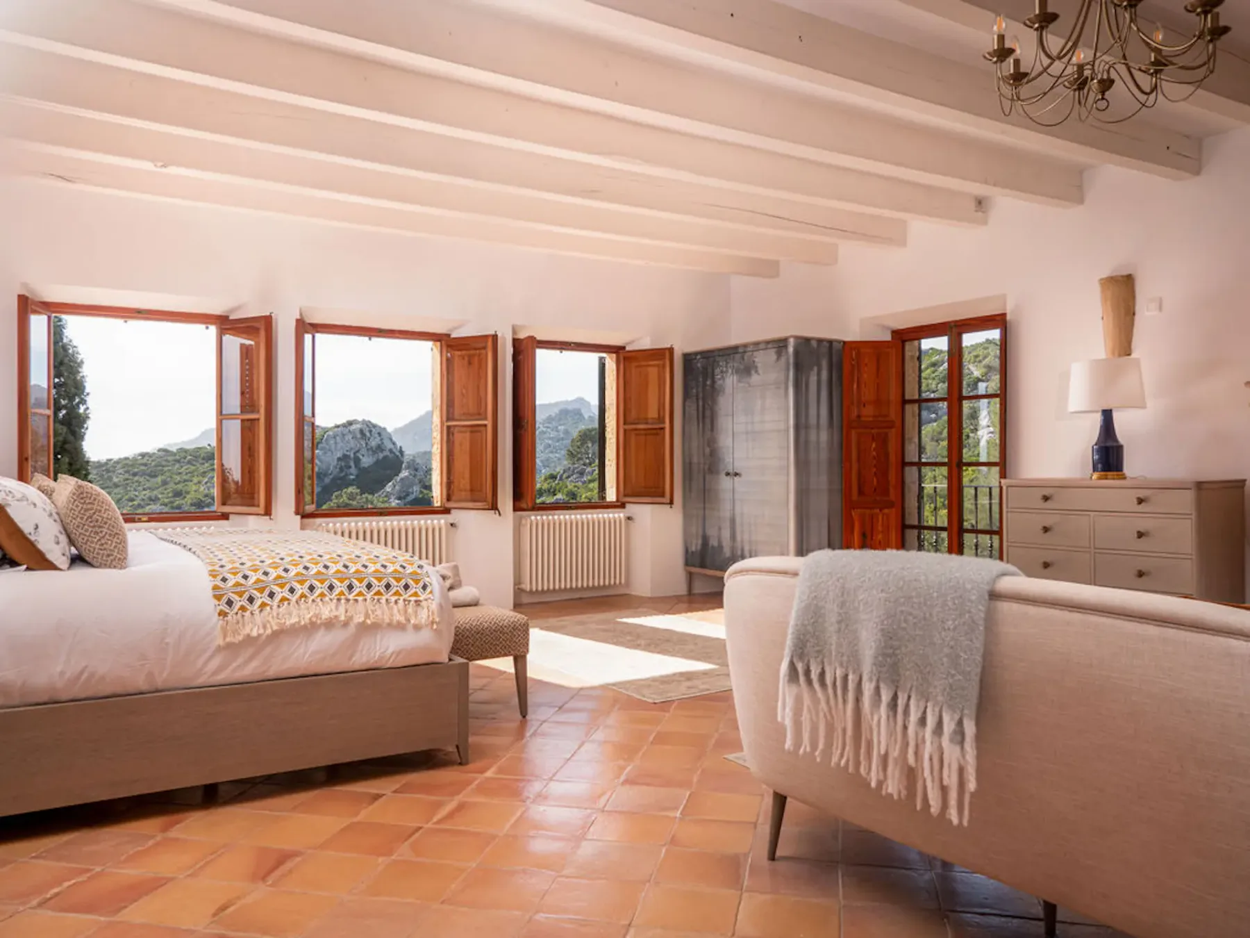 A bedroom with a bed, seating, and open windows overlooking the hills.