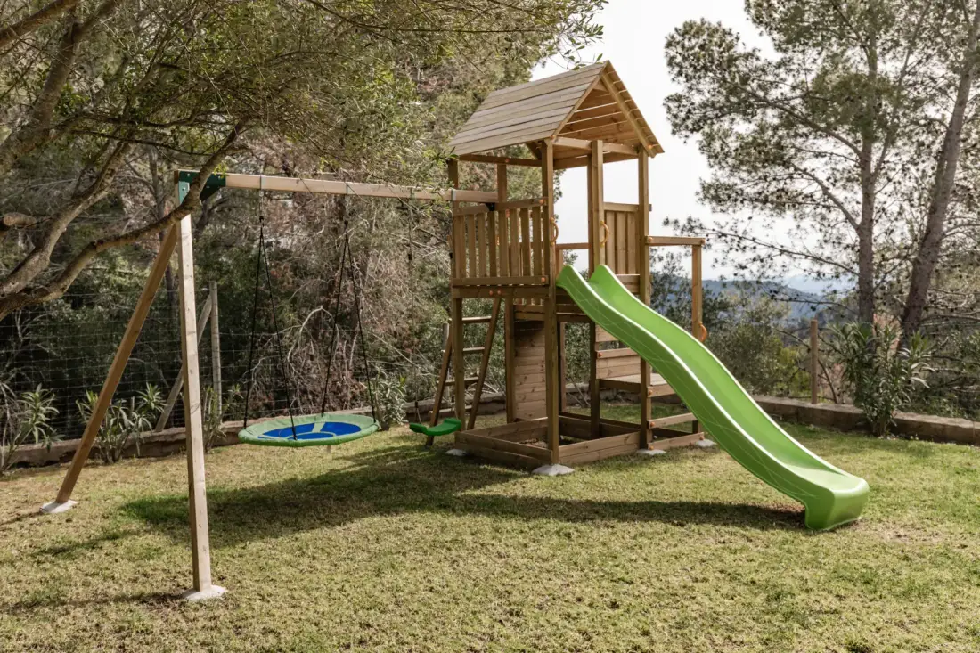 A wooden children’s play structure with a green slide stands in a grassy yard.