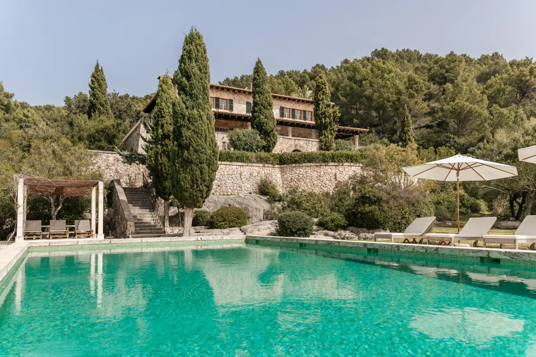 A turquoise swimming pool sits below a hillside building and surrounding trees.