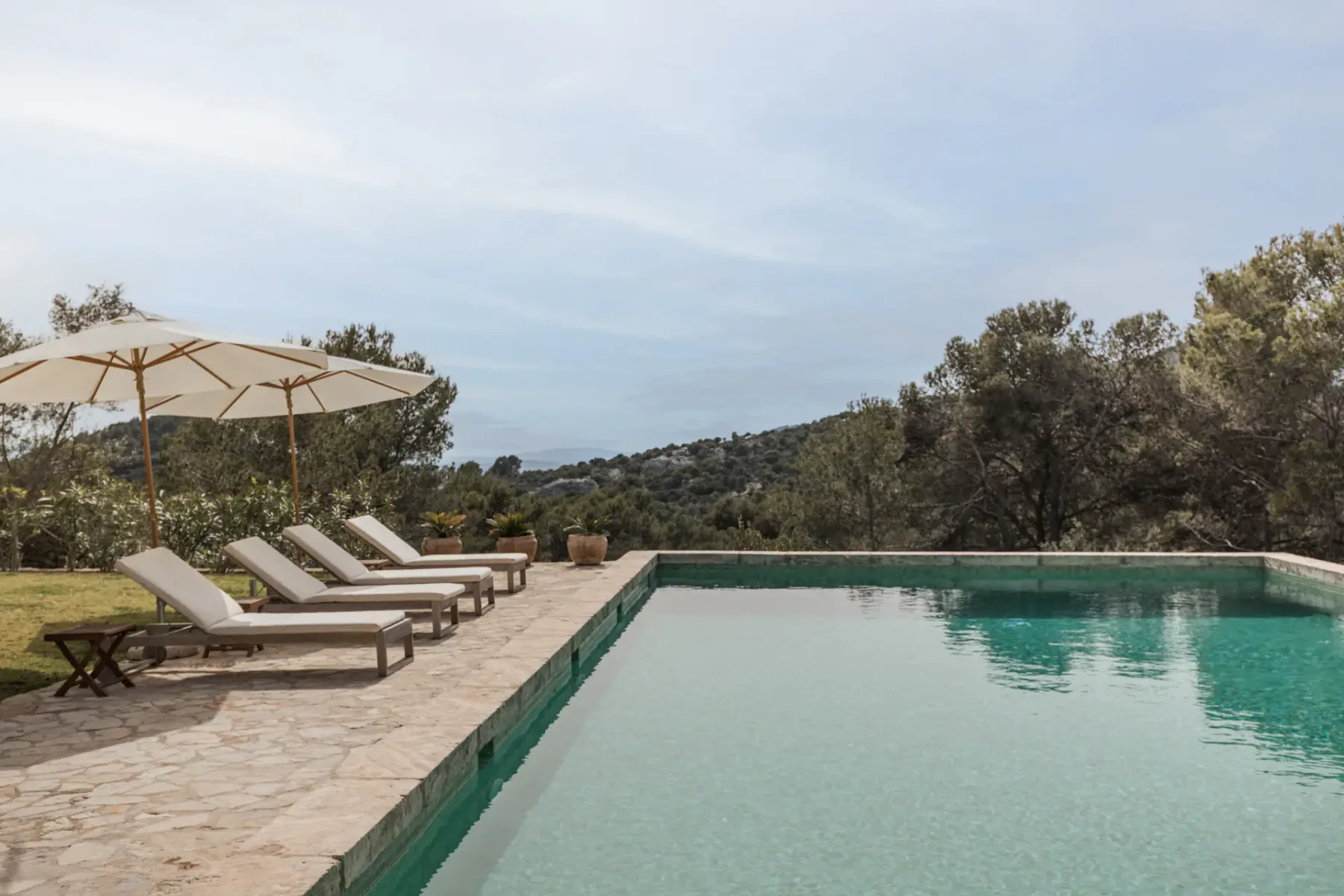 An outdoor swimming pool beside sun loungers, with wooded hills in the background.