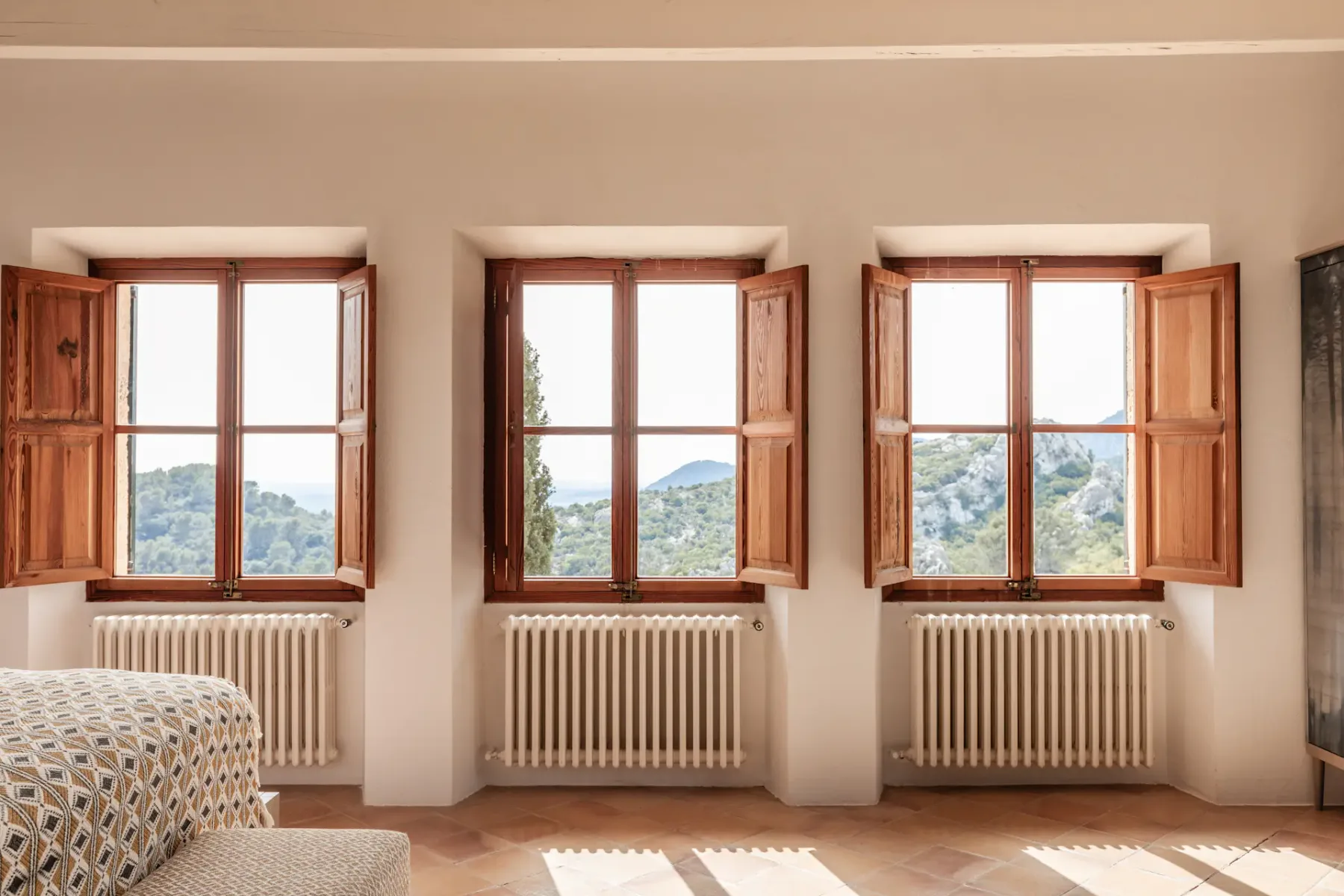 A bedroom with patterned bedding beside three windows overlooking green hills.