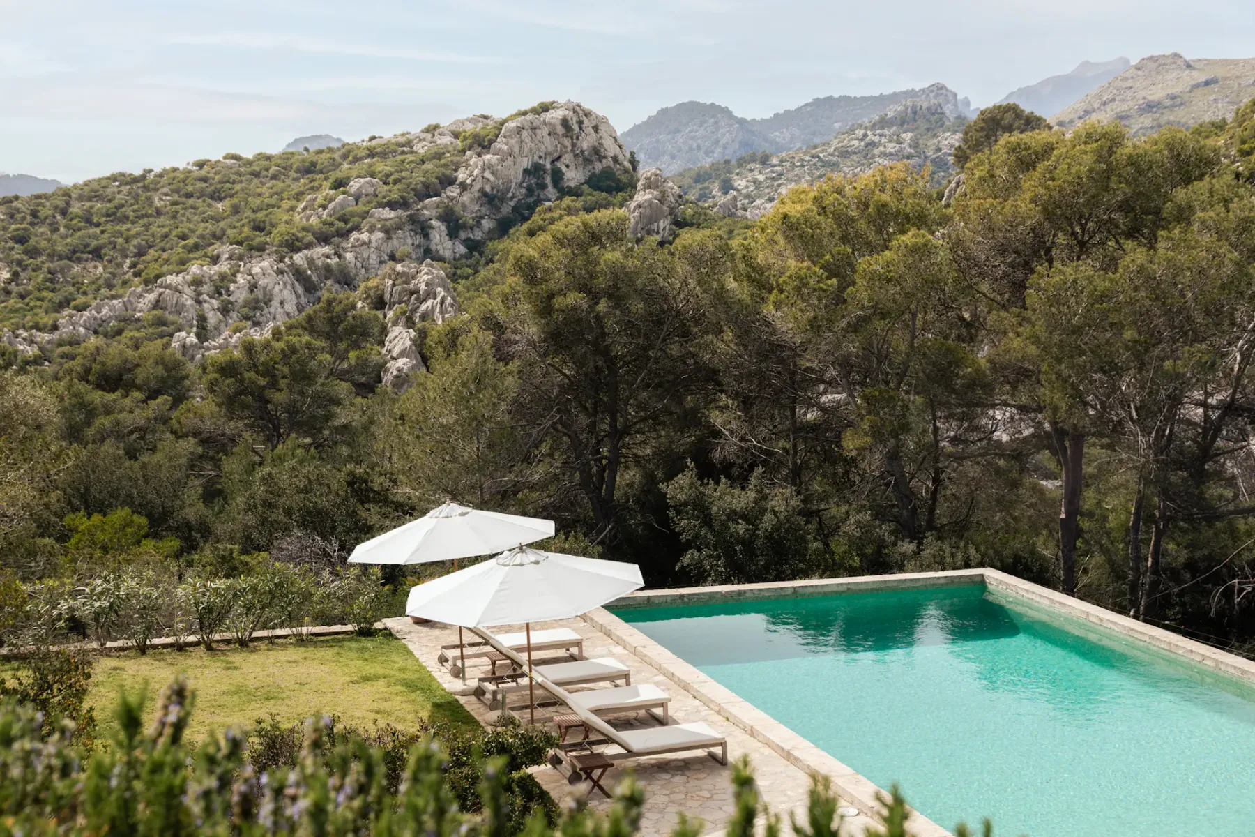 A swimming pool and loungers sit beside a green hillside with mountains beyond.
