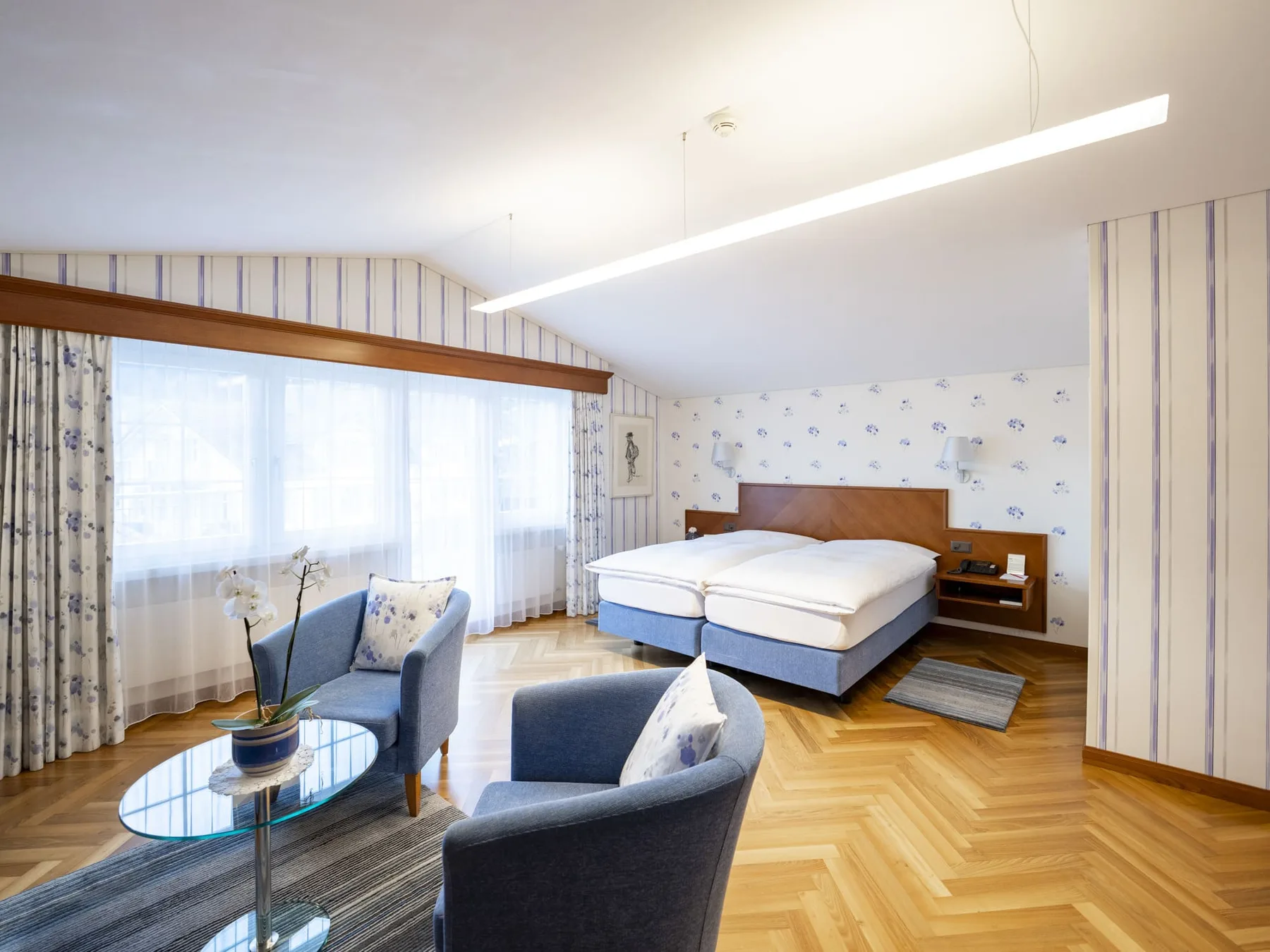Hotel room with parquet flooring, blue chairs, a double bed and large windows.