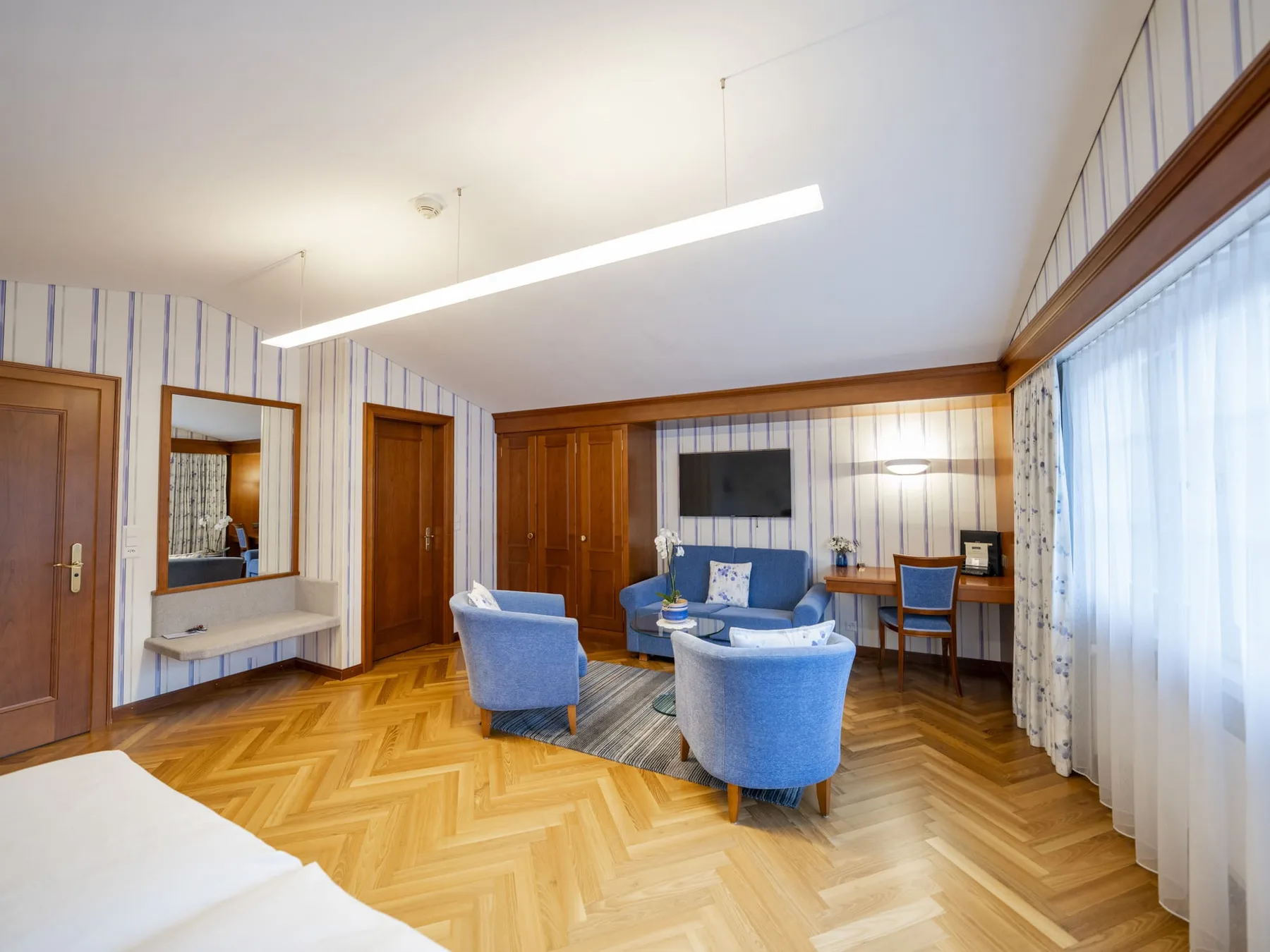 Panorama Junior Suite sitting area with a blue sofa, TV area, desk and wooden wardrobes.