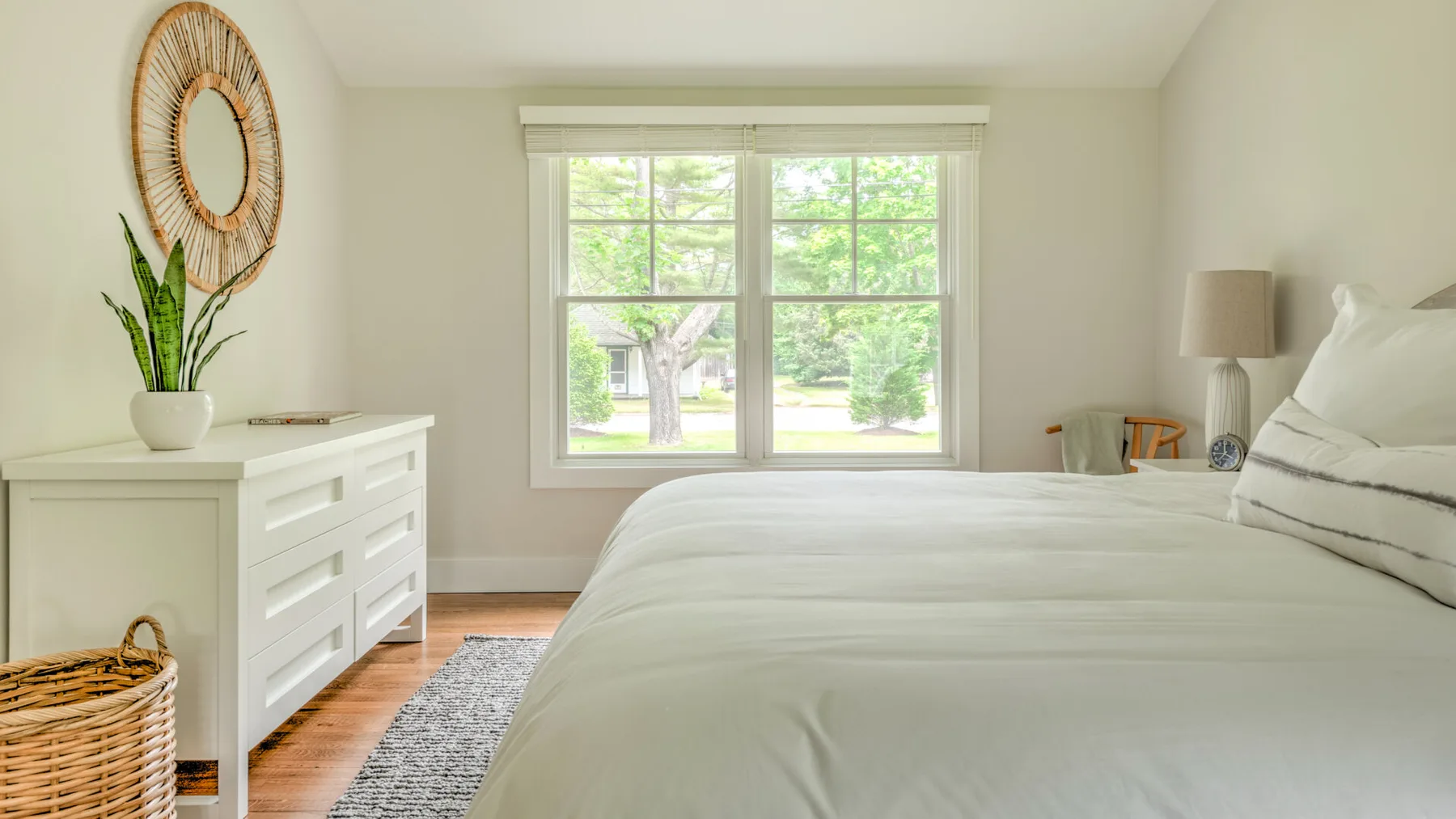 A bright bedroom with a large bed, white dresser, and window overlooking greenery.