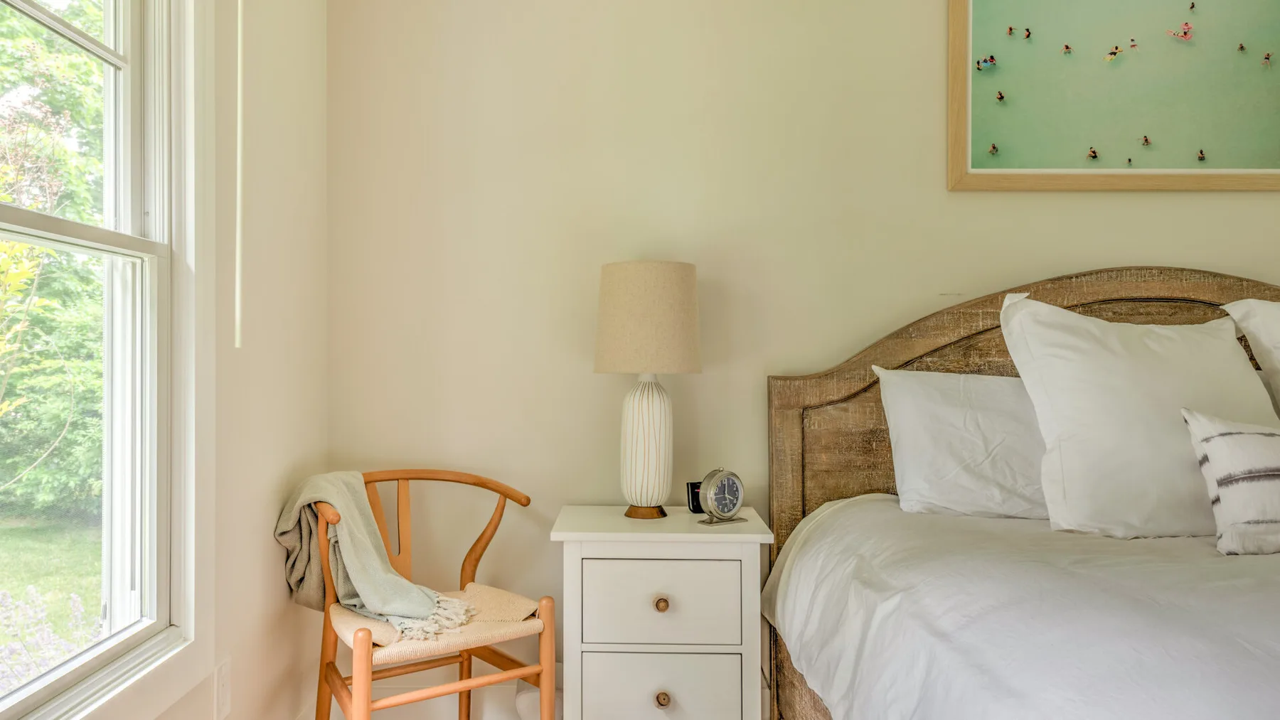 A bright bedroom with a bed, bedside table, and a chair beside the window.