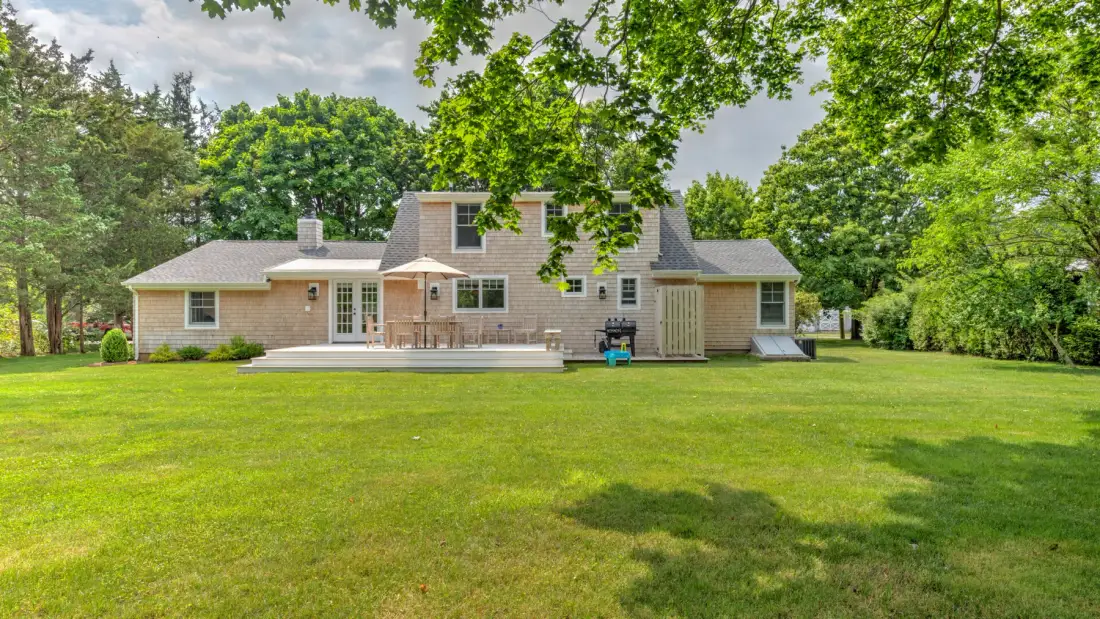 A house stands beyond a broad green lawn, surrounded by mature trees.