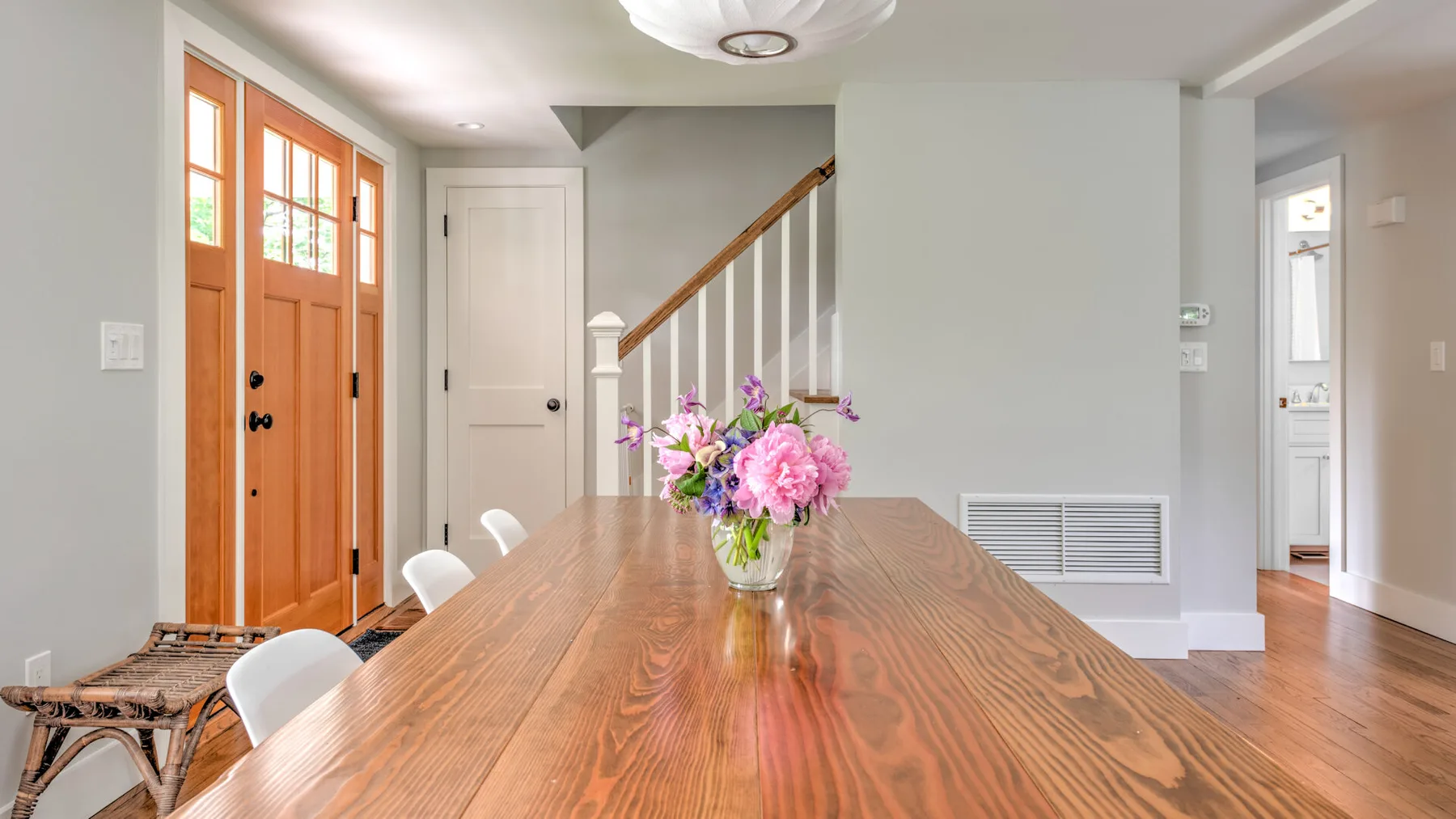 A wooden dining table with flowers sits in a bright room beside a staircase.