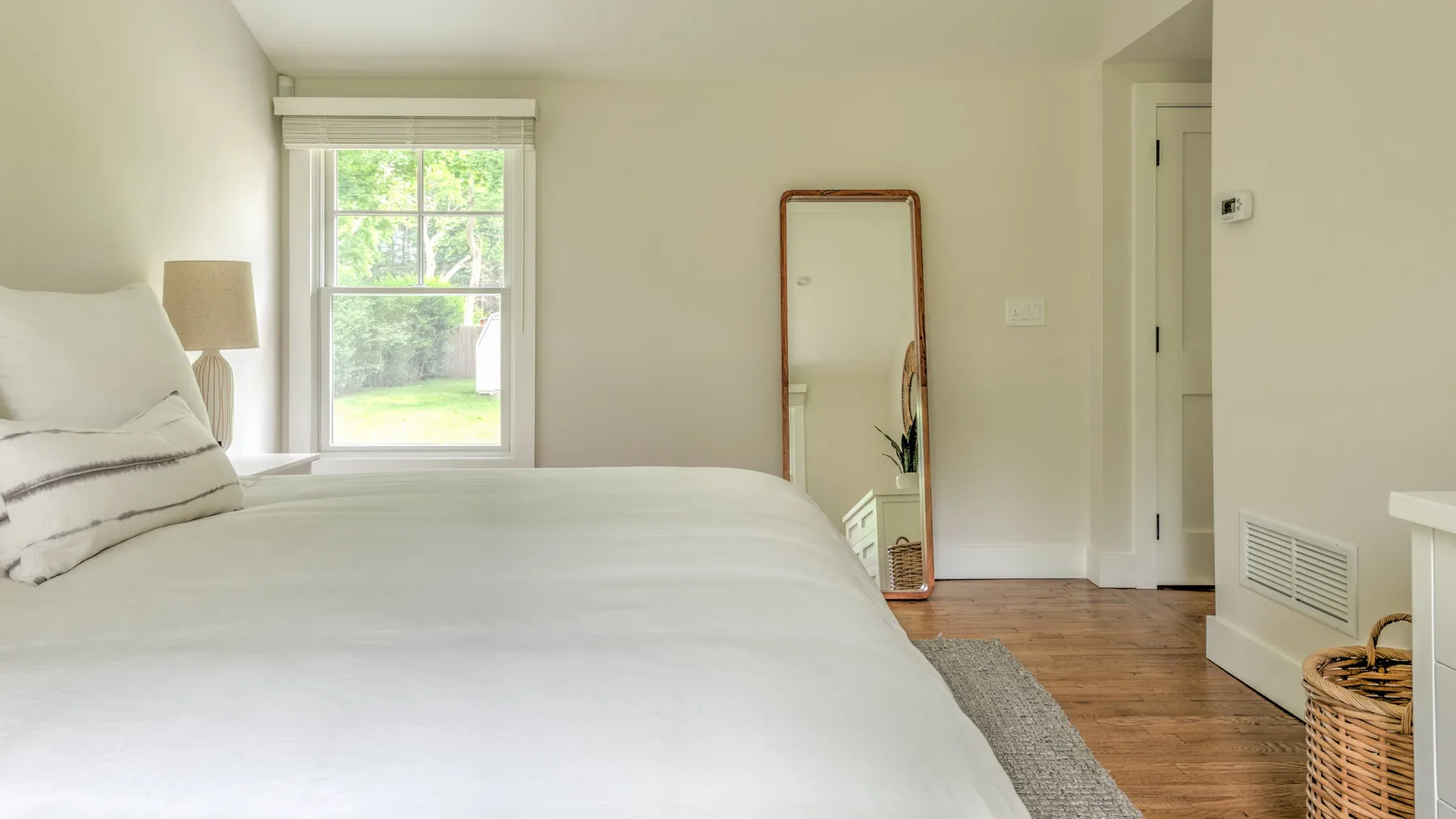 A bright bedroom with a white bed beside a window and a standing mirror.