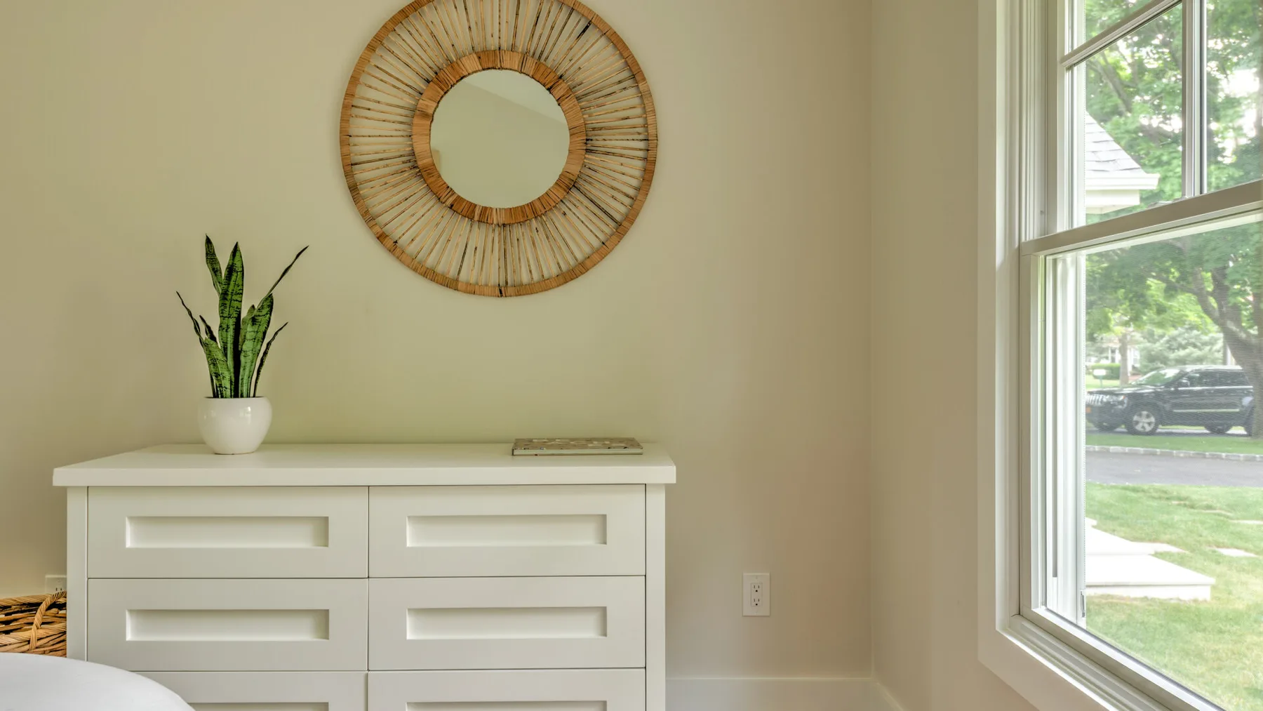 A bright room with a white dresser, round mirror, and window.