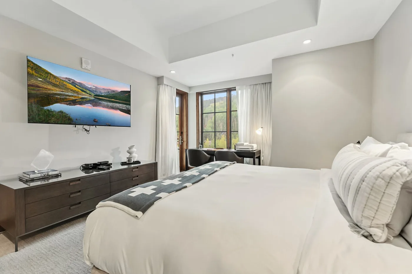 A bright bedroom with a large bed and a television mounted above a low dresser.