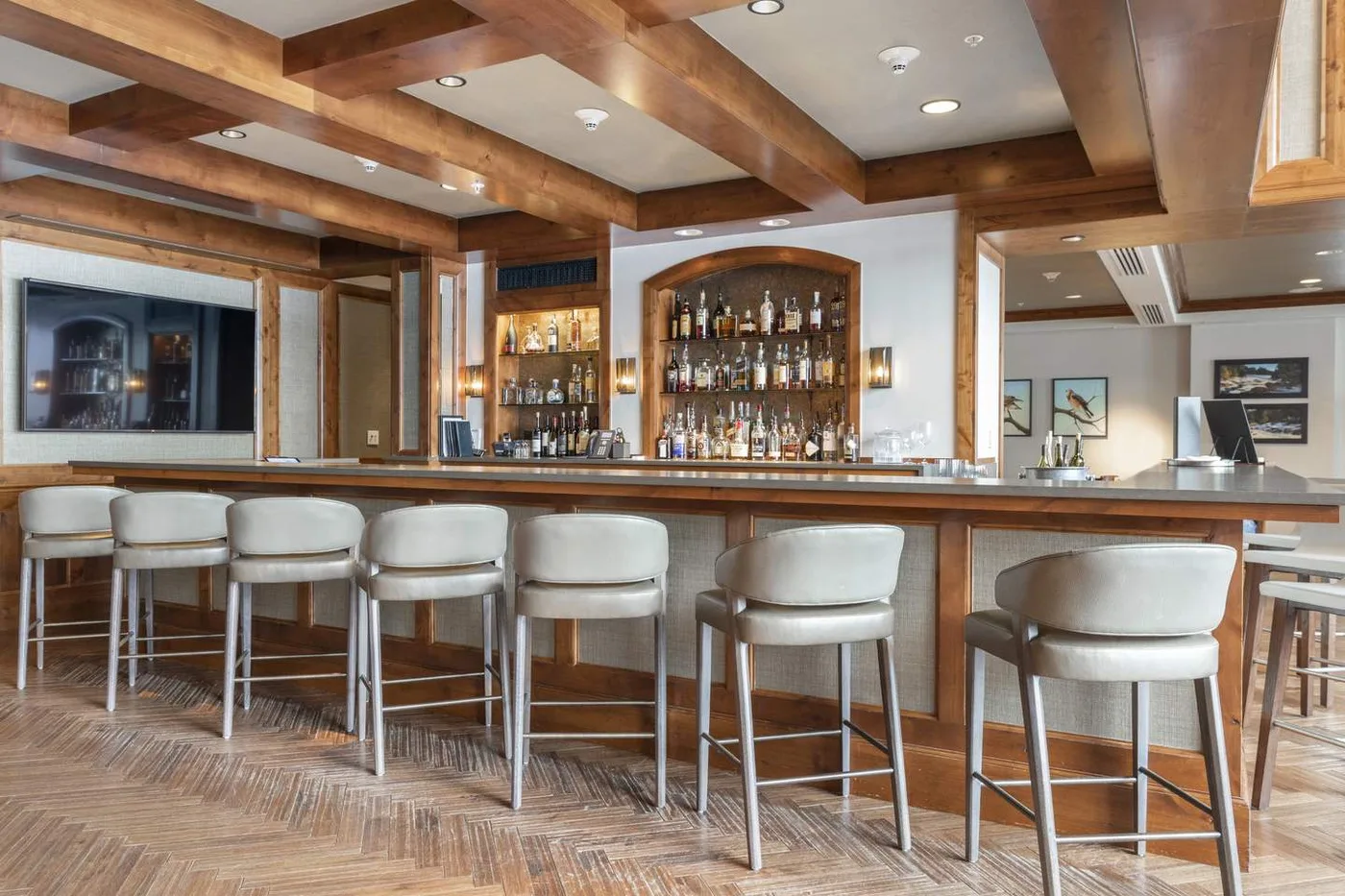 A bar counter with stools and shelves of bottles beneath exposed wooden beams.