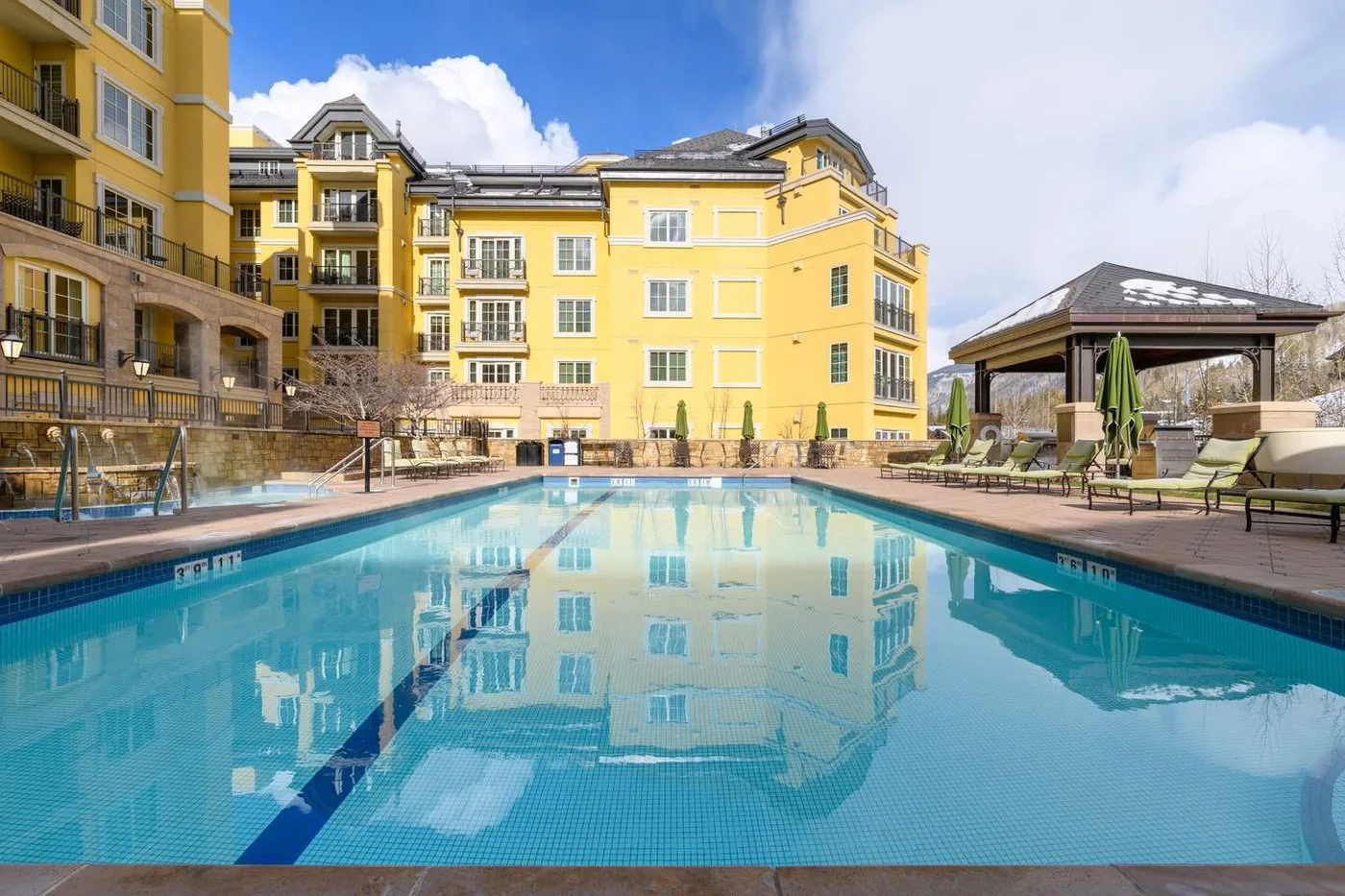 An outdoor swimming pool sits in front of yellow multi-story buildings and a shaded seating area.