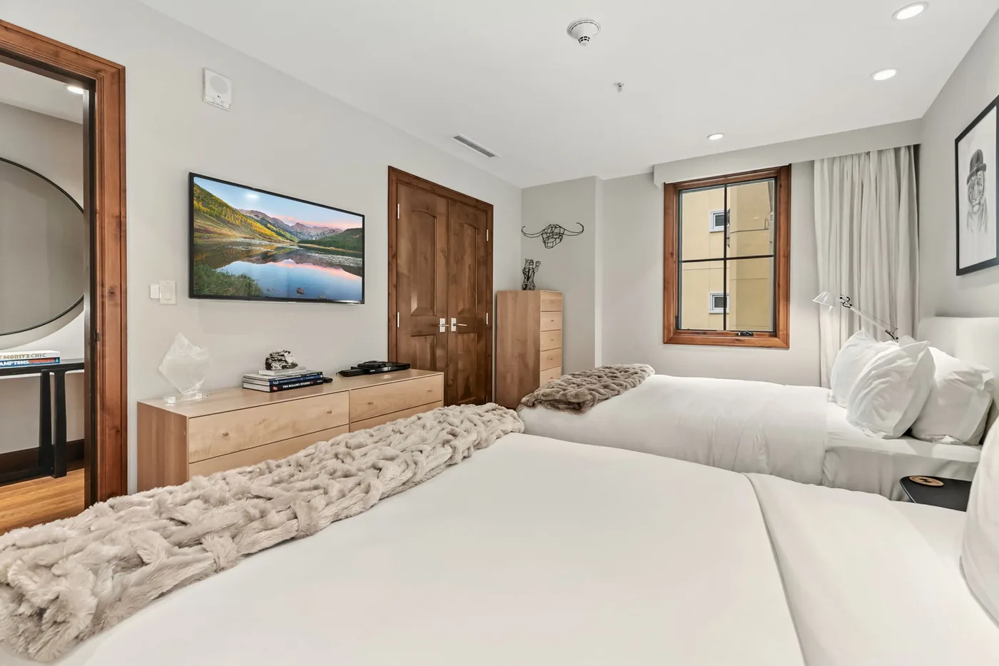 A bright bedroom with two beds, a wooden dresser, and a window with curtains.