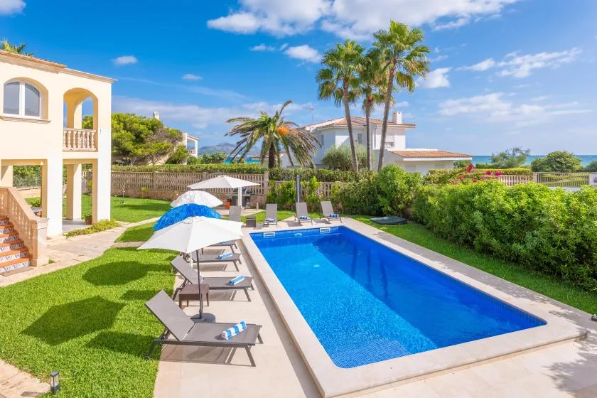Una piscina exterior junto a un patio soleado y un jardín ajardinado.