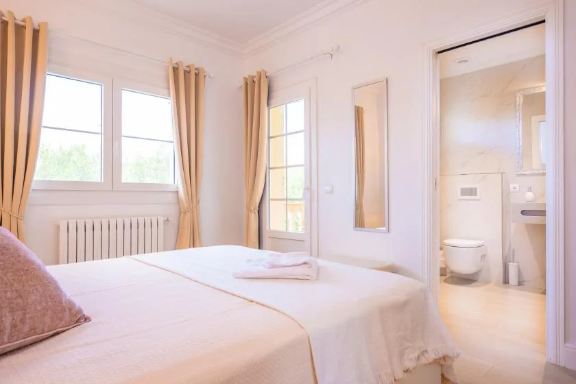 Un dormitorio luminoso con una cama hecha junto a las ventanas y la puerta abierta del baño.