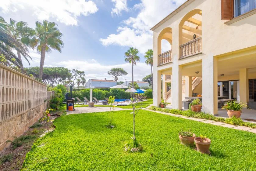 Un patio con césped junto a un edificio color crema, con palmeras y una piscina al fondo.