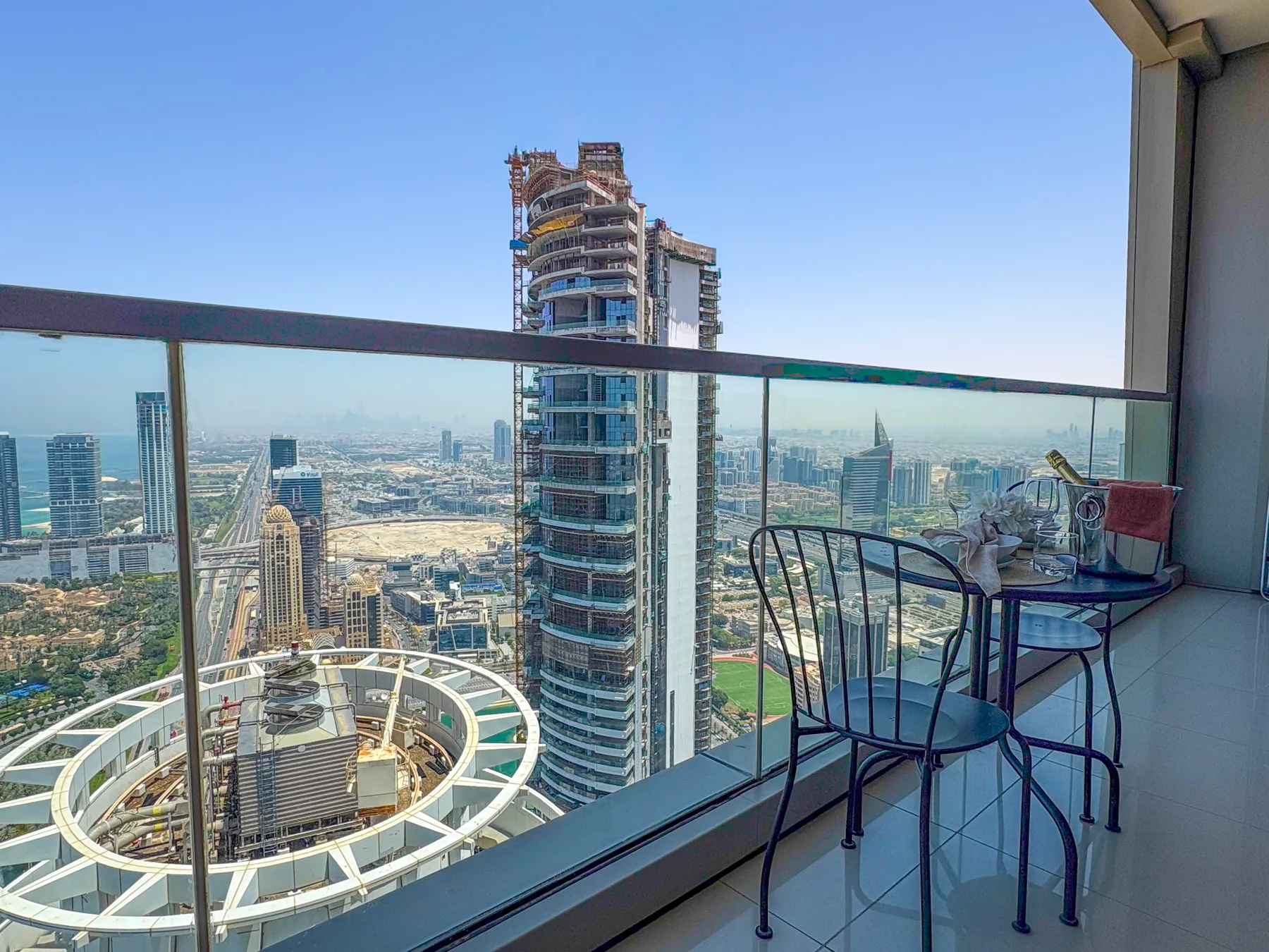 A balcony overlooks a city skyline, with a glass railing and outdoor seating.
