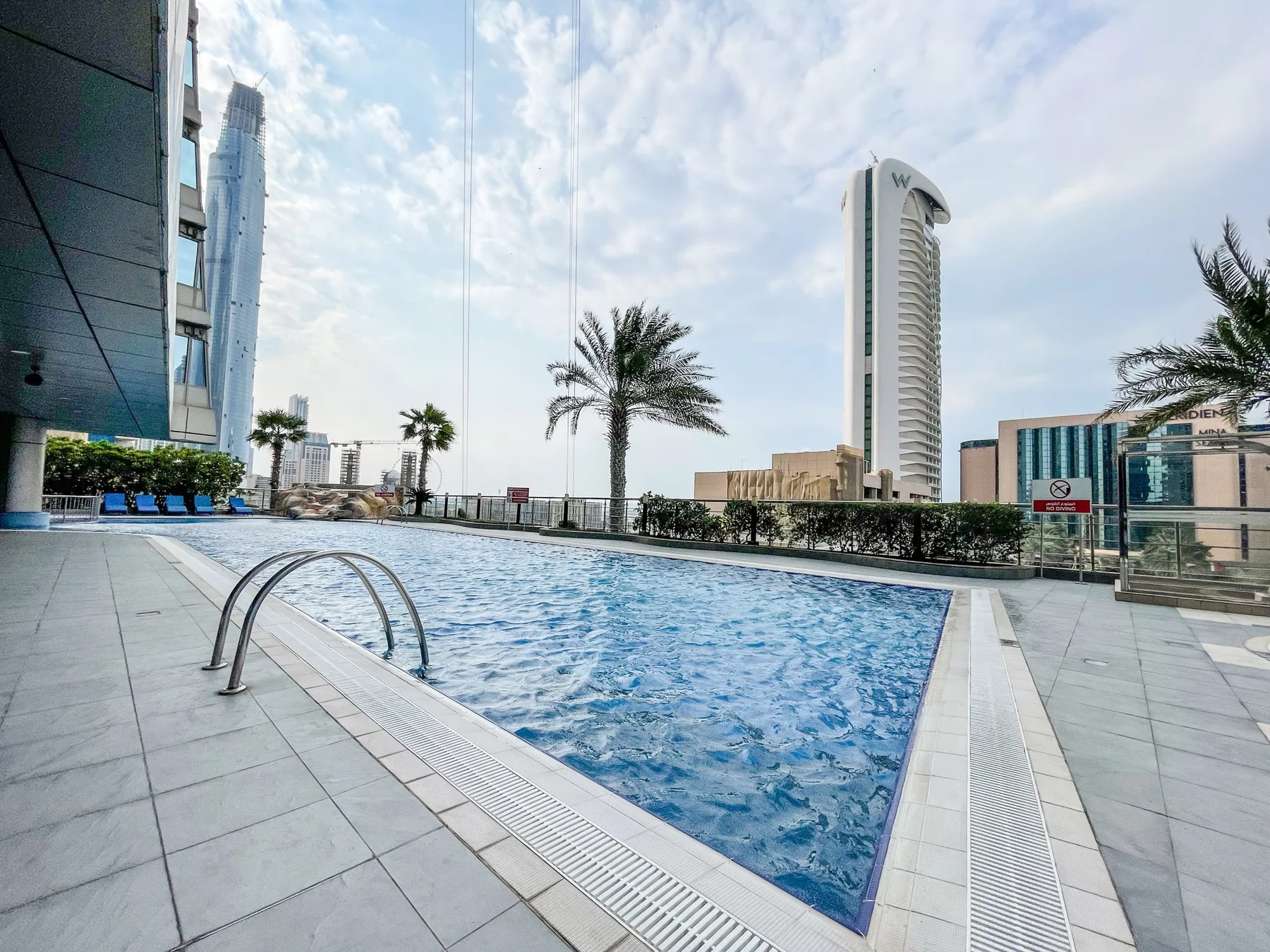 A rooftop swimming pool sits beside a tiled deck with palm trees and city buildings beyond.