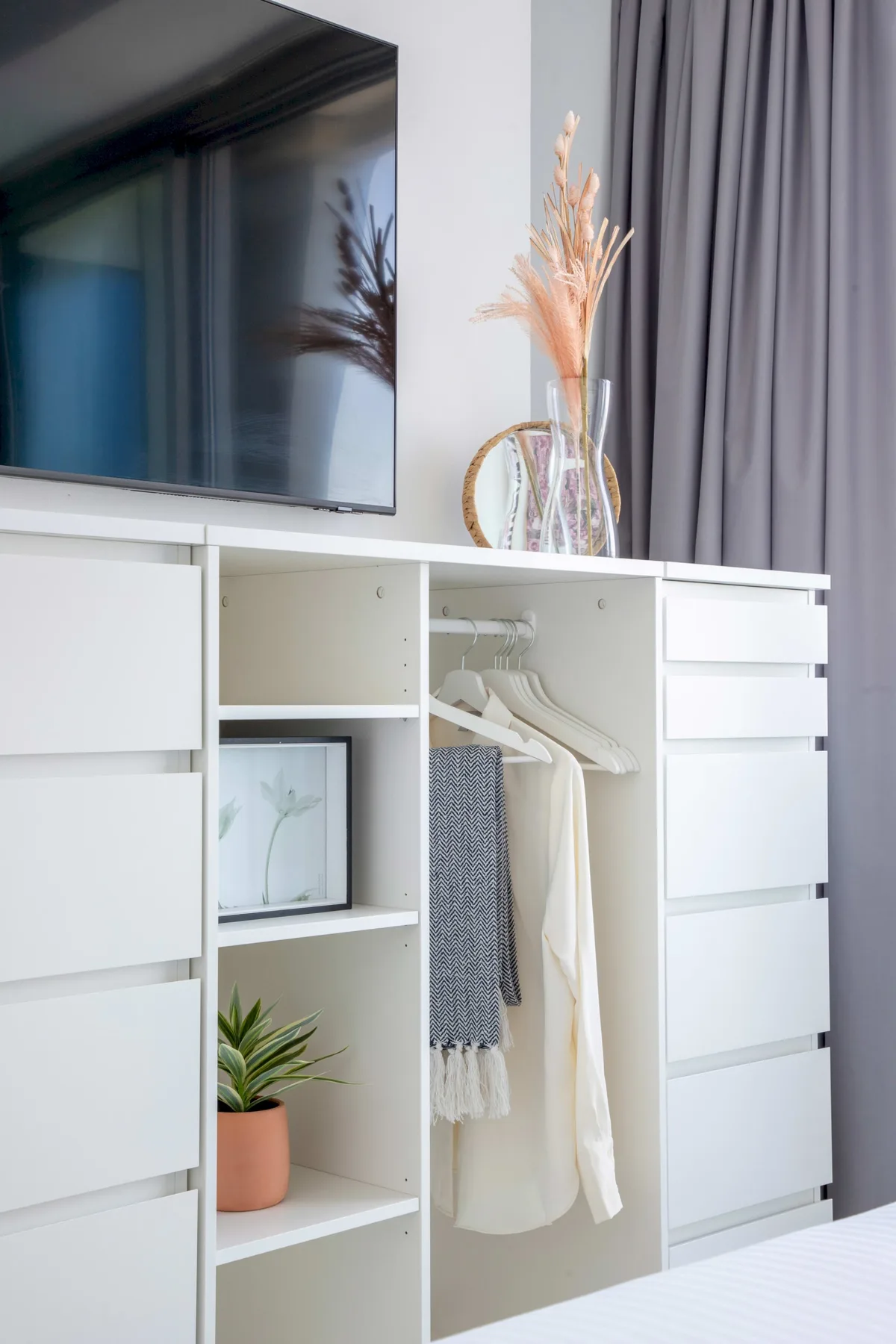 A white bedroom storage unit sits beneath a wall-mounted television, with curtains behind it.