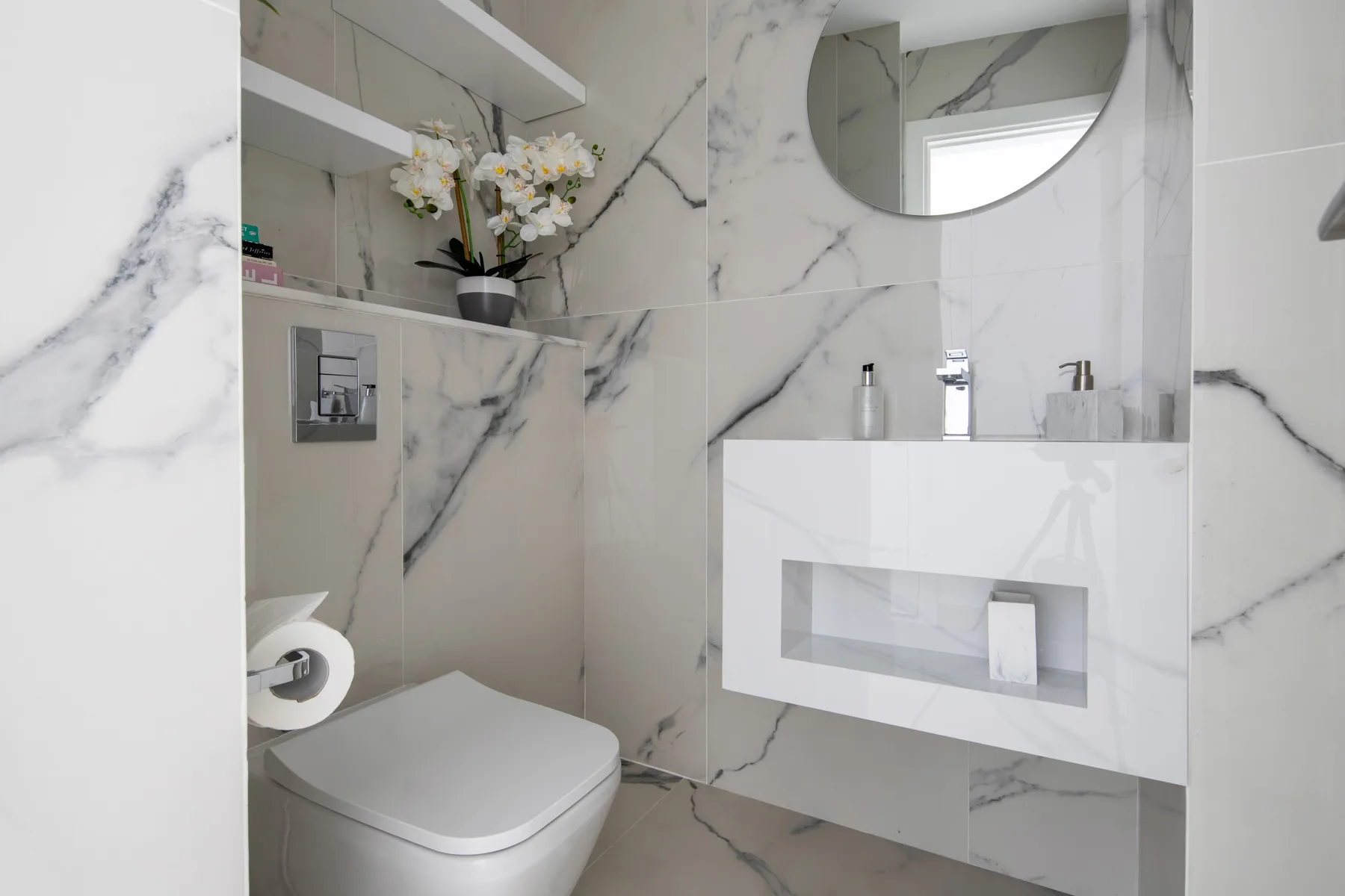 A marble-look bathroom with a toilet, wall-mounted sink, and round mirror.