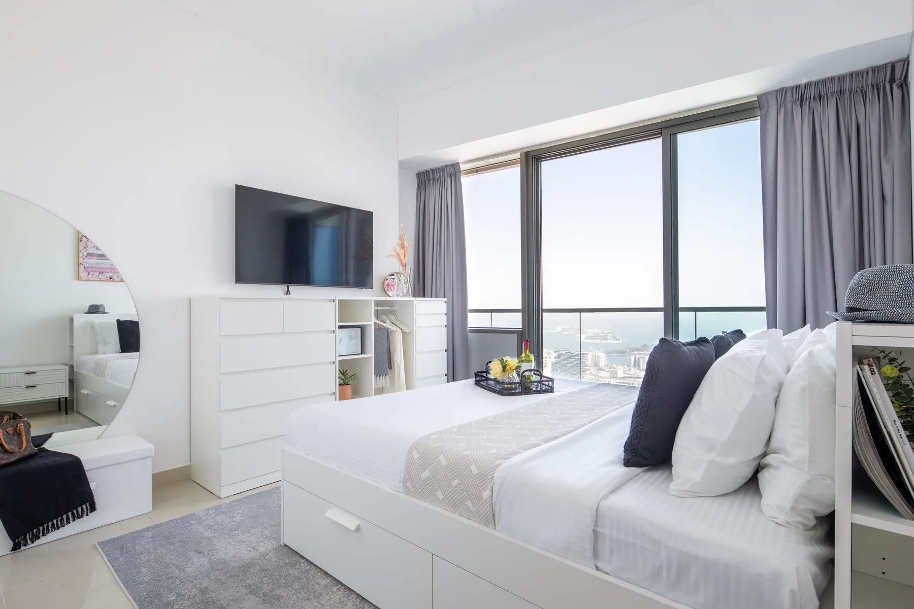 A bright bedroom with a neatly made bed beside large windows overlooking the city.