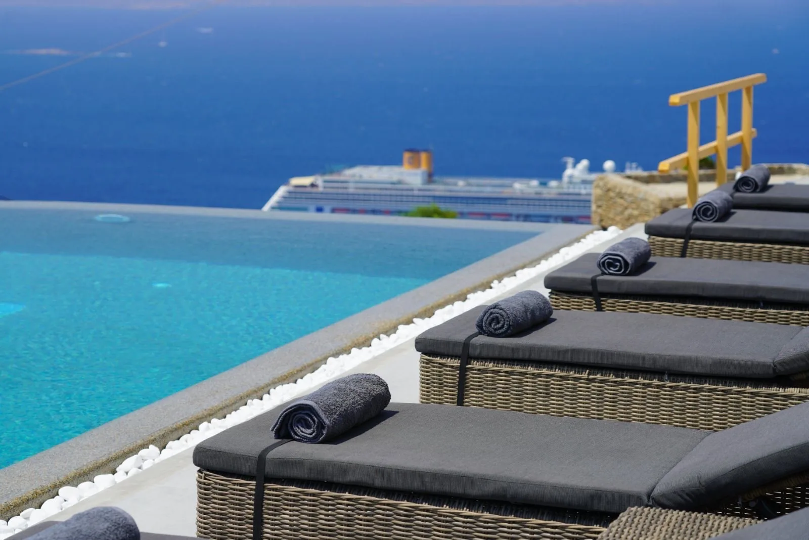 Poolside loungers overlook the sea, with a cruise ship visible in the distance.