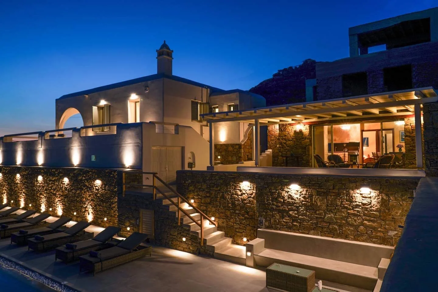 A modern villa glows at dusk above a stone terrace with outdoor seating.