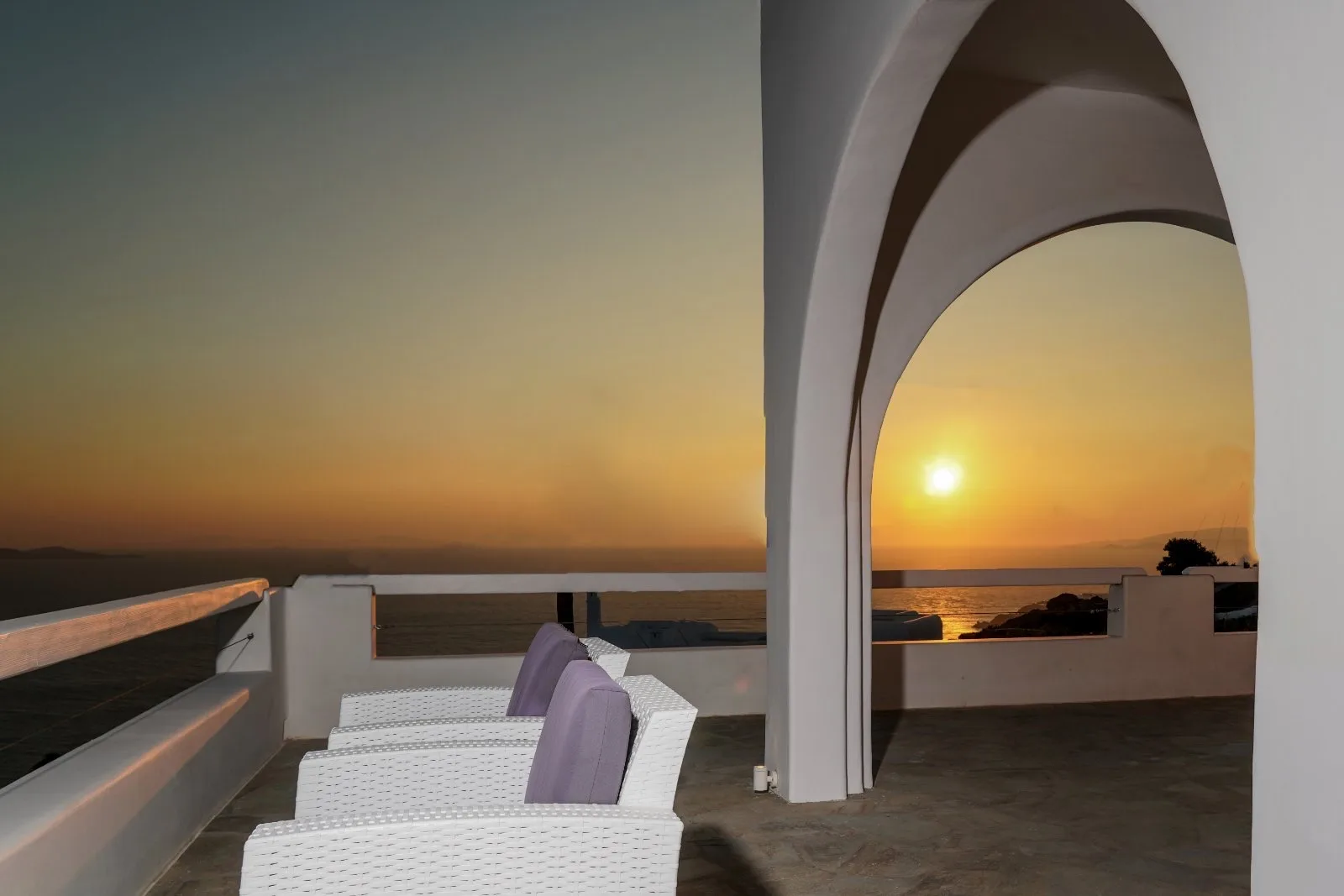 A rooftop terrace with white seating overlooks the ocean at sunset.