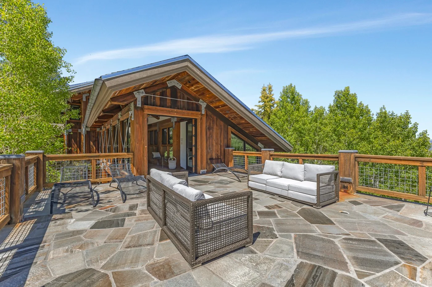 A wooden cabin opens onto a spacious stone terrace with outdoor seating and trees beyond.