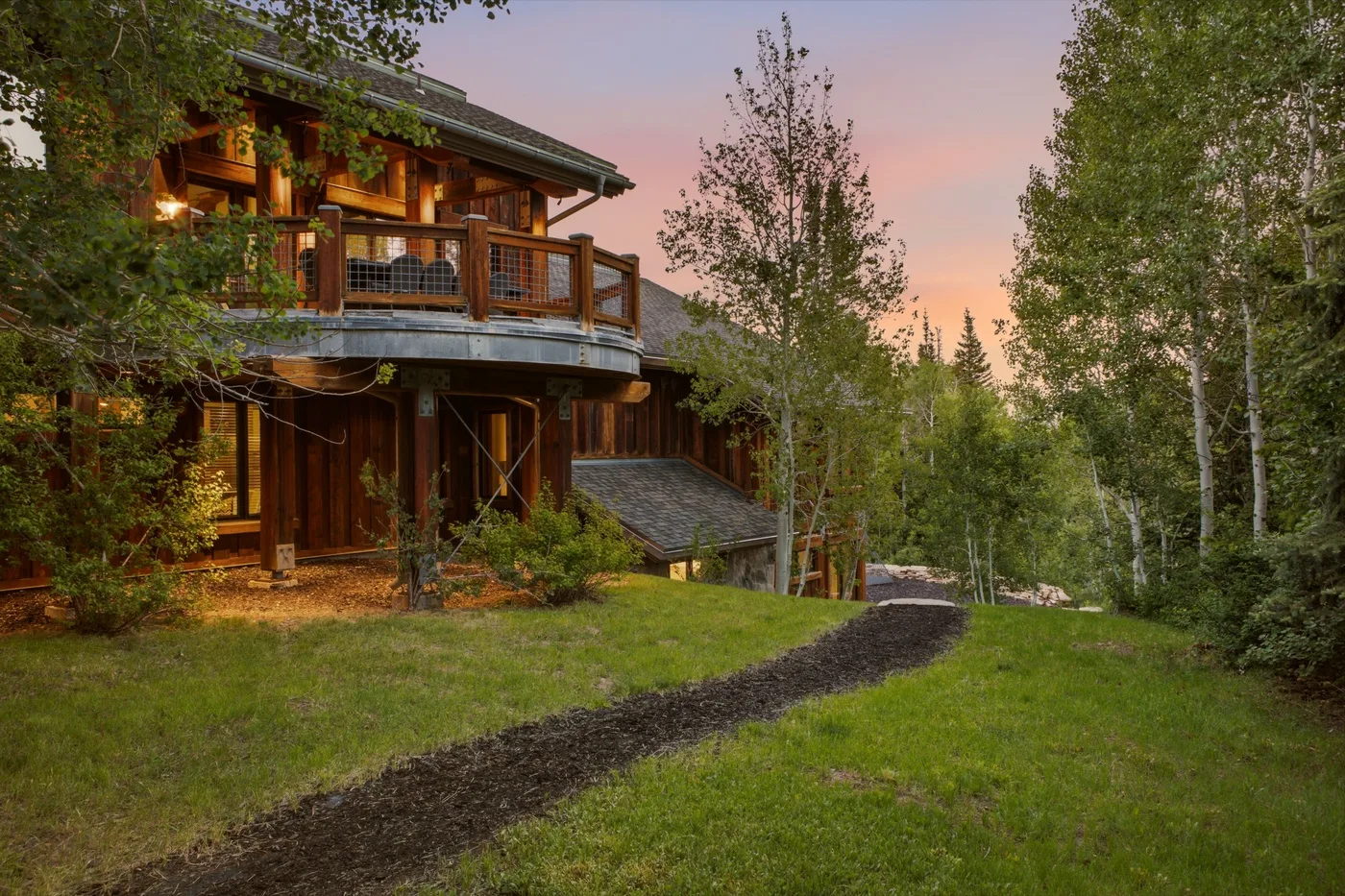 A wooden house with a balcony stands beside a grassy yard at sunset.