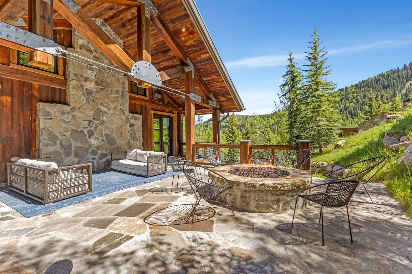 A stone patio beside a timber lodge features outdoor seating and a circular fire pit.