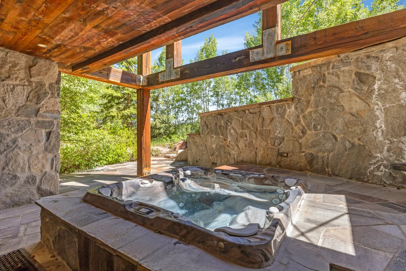 An outdoor hot tub sits beneath a timber roof beside stone walls.