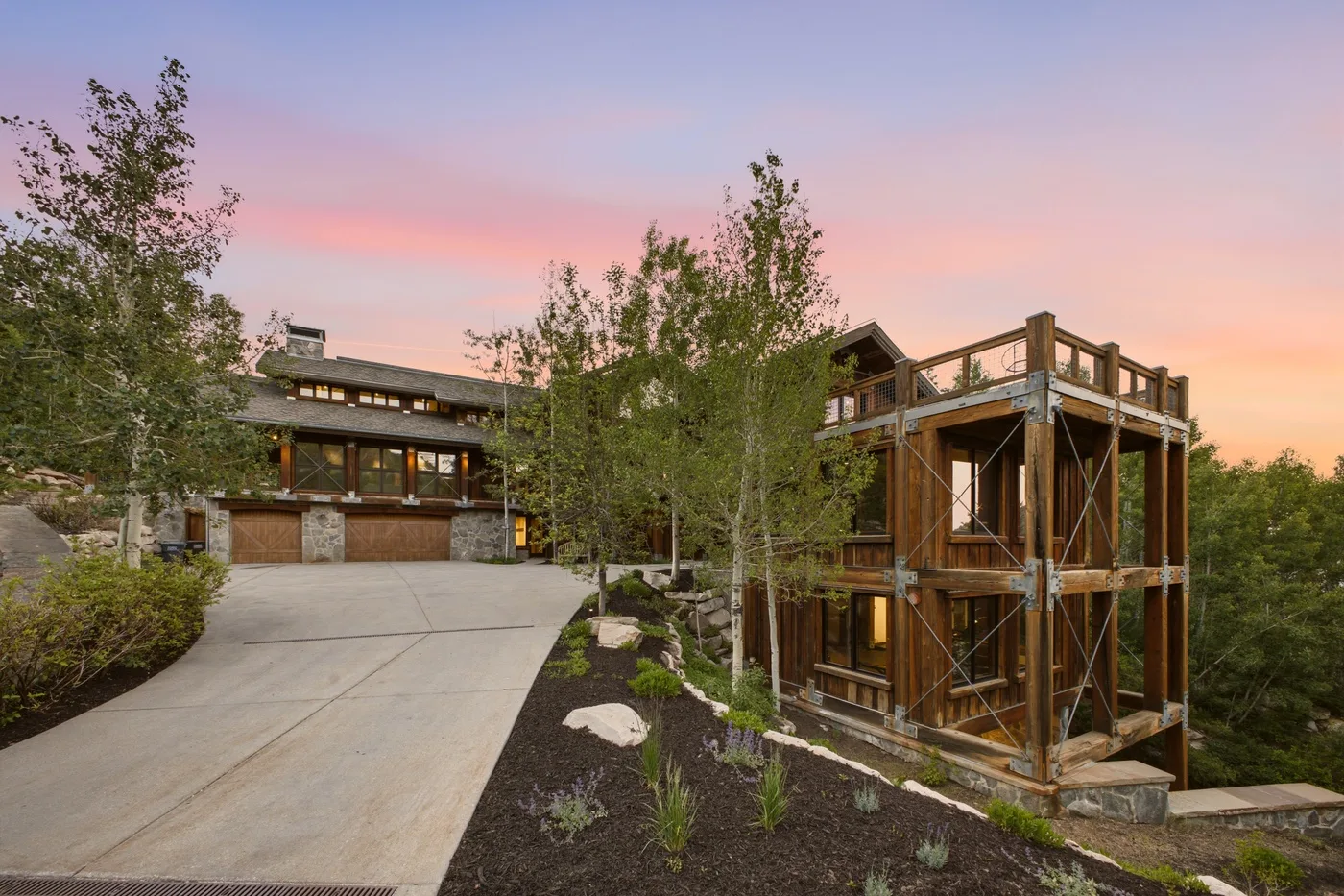 A modern house and a timber-framed building sit beside a sloping driveway at sunset.