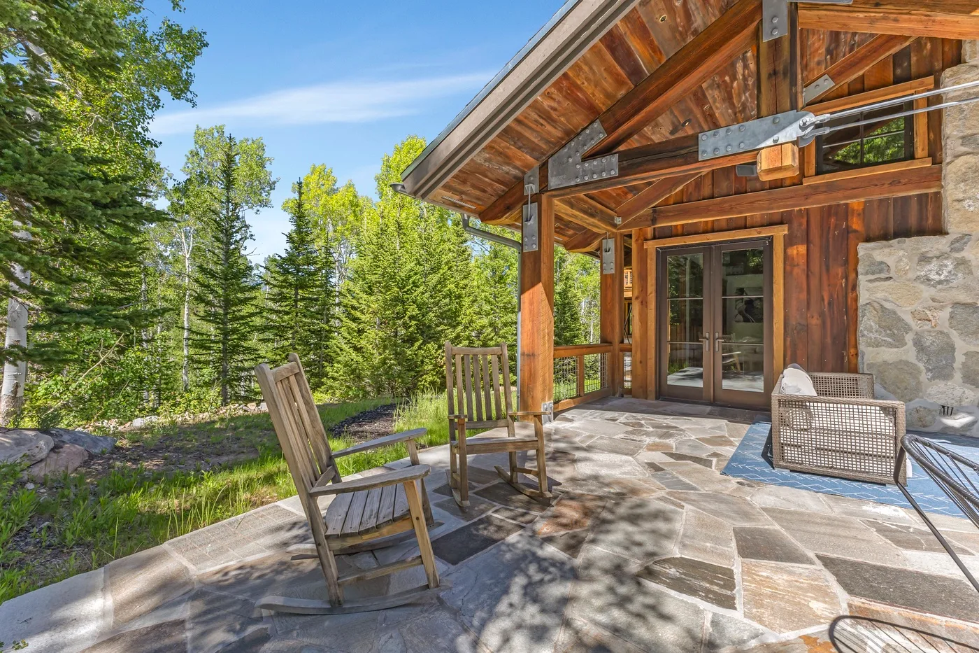 A covered stone patio beside a timber-sided house, with outdoor seating and trees.