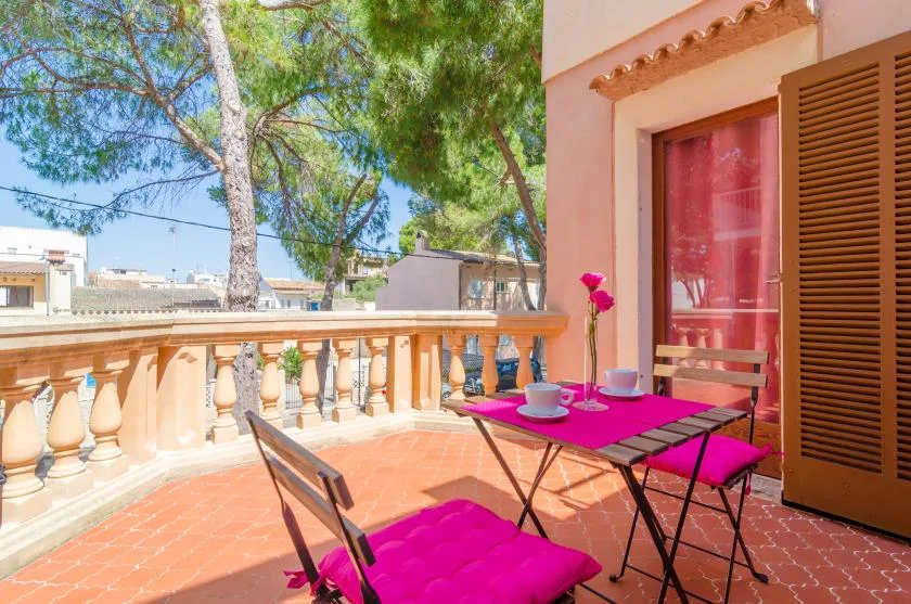 A sunny terrace with a pink-covered table and chairs beside an open shuttered doorway.
