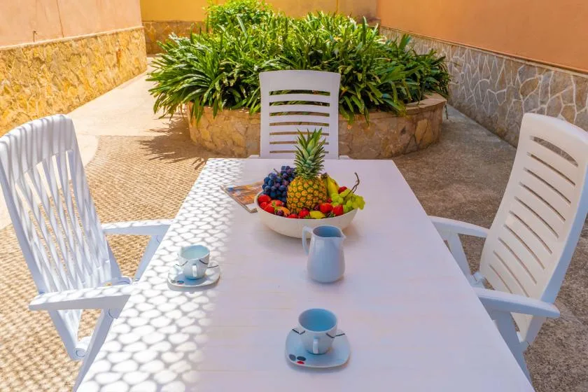 An outdoor patio table is set with fruit and cups beside a leafy planter.