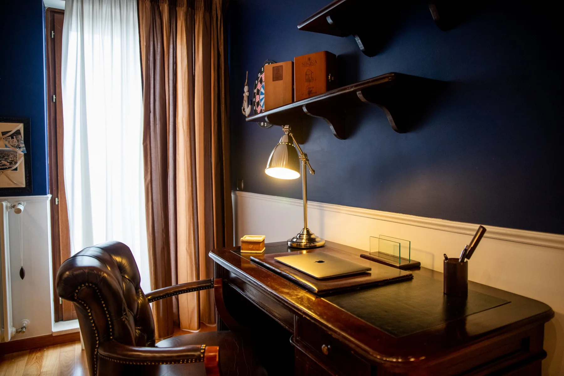 A study desk sits beside a tall window with curtains in a dark blue room.