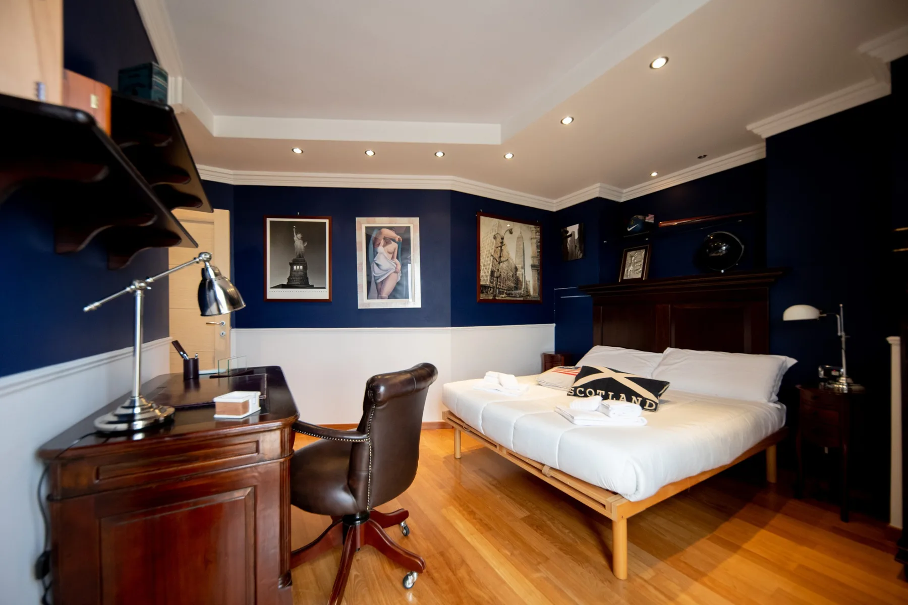 A bedroom with a large bed, wooden desk, and framed artwork on dark blue walls.