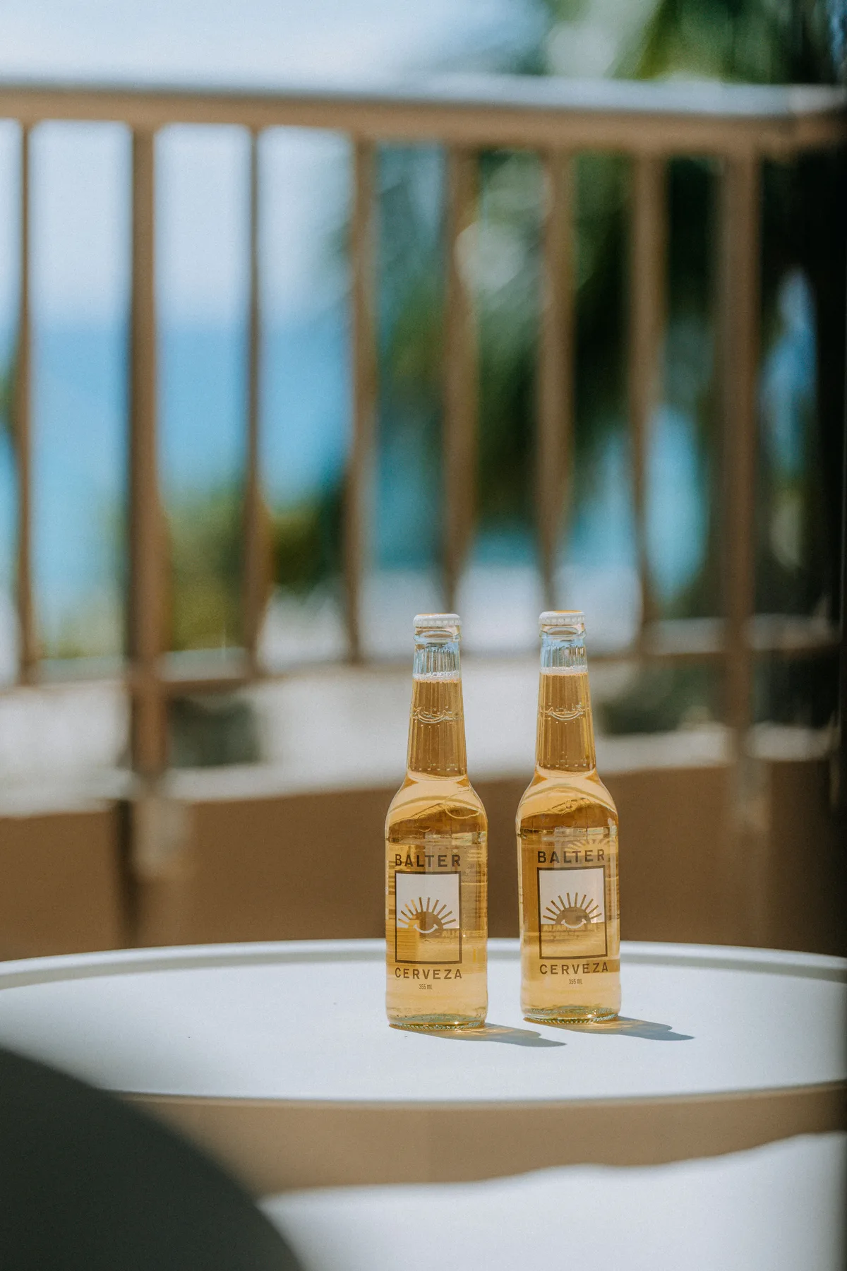Two bottles sit on a round table beside a balcony railing overlooking the ocean.