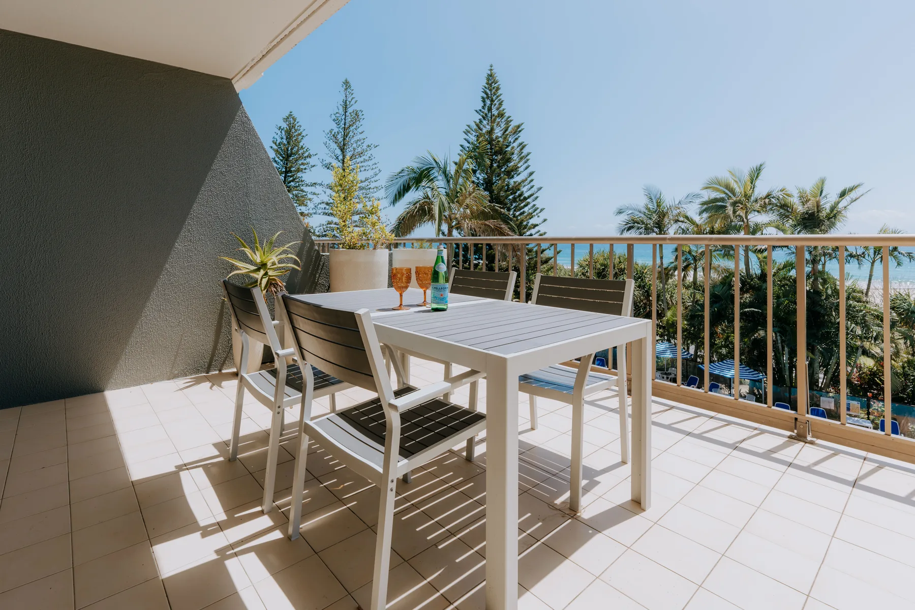 A sunlit balcony has a white table and seating beside a railing.