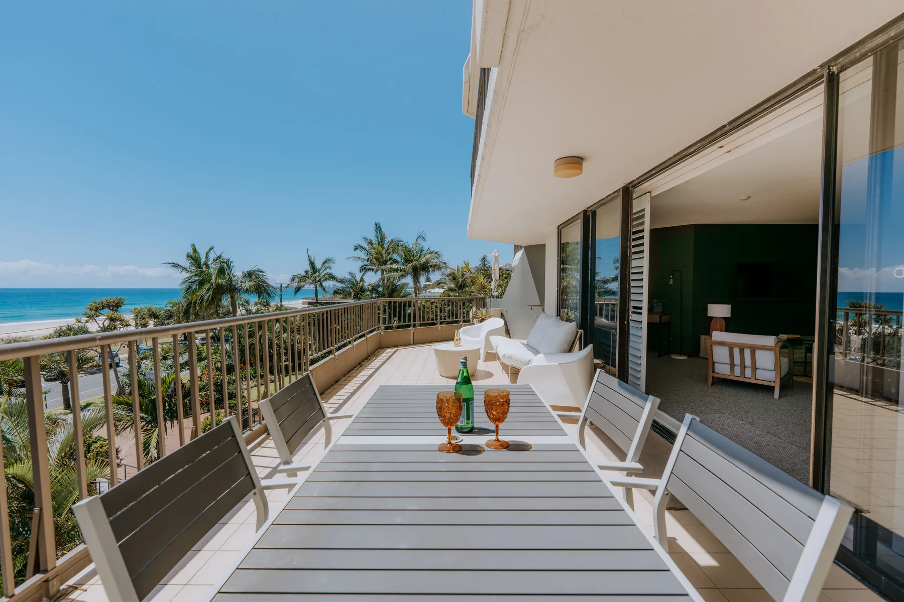 A balcony with outdoor seating overlooks palm trees and the ocean.