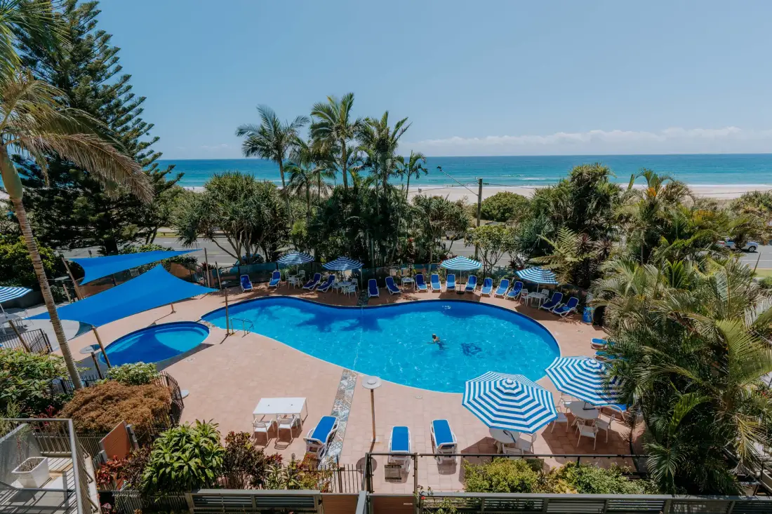 A resort pool area with blue umbrellas, palm trees, and the ocean beyond.