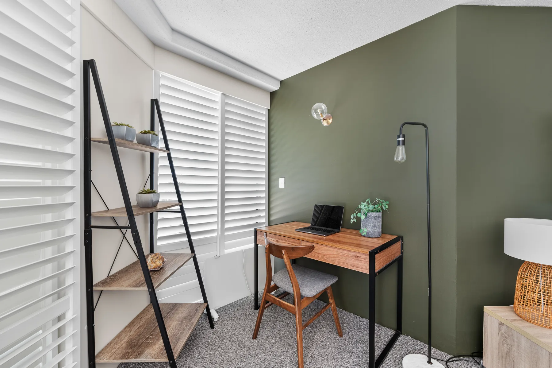 A compact workspace with a wooden desk and shelving beside shuttered windows.
