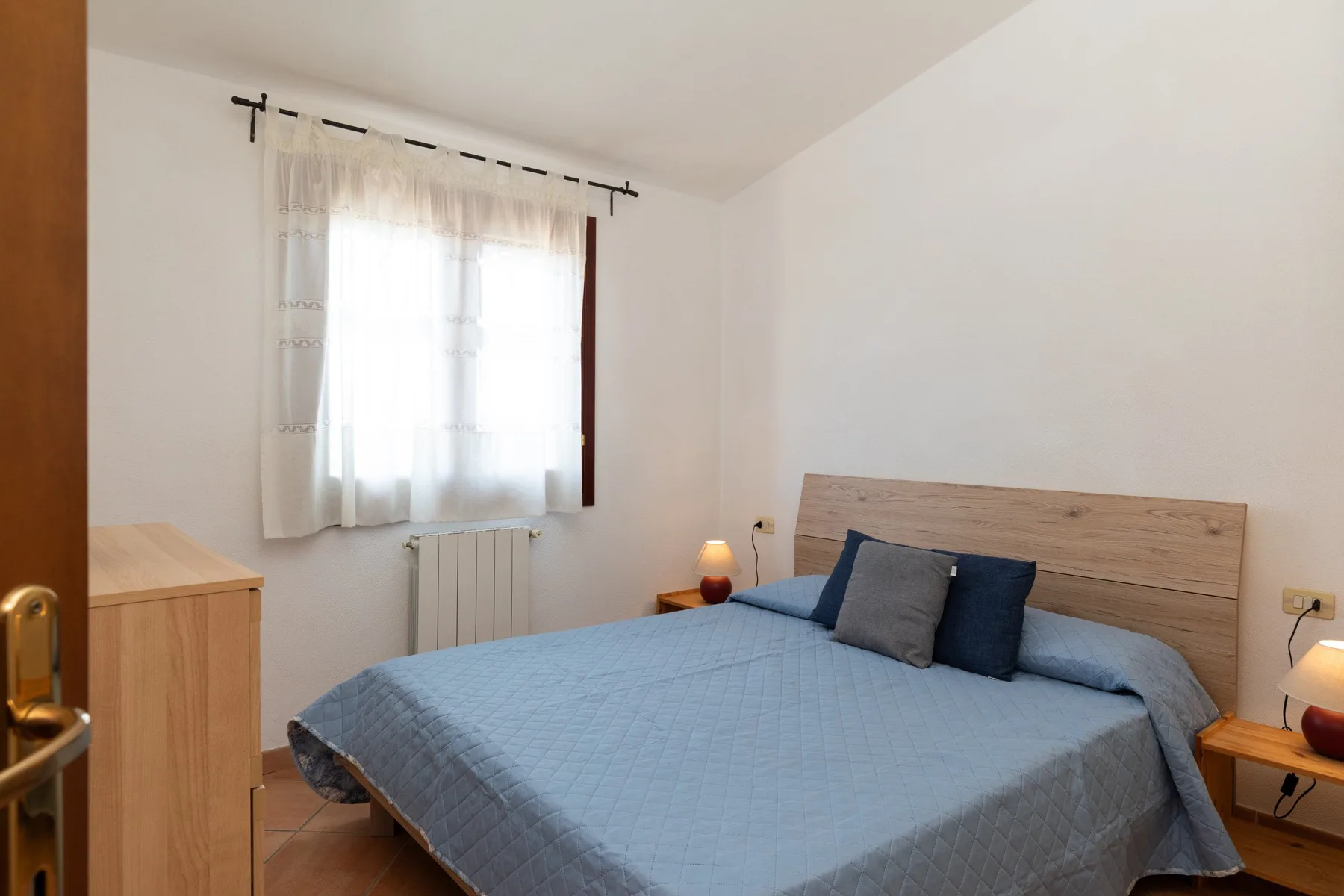 Un dormitorio con una cama bajo una ventana con cortinas translúcidas.