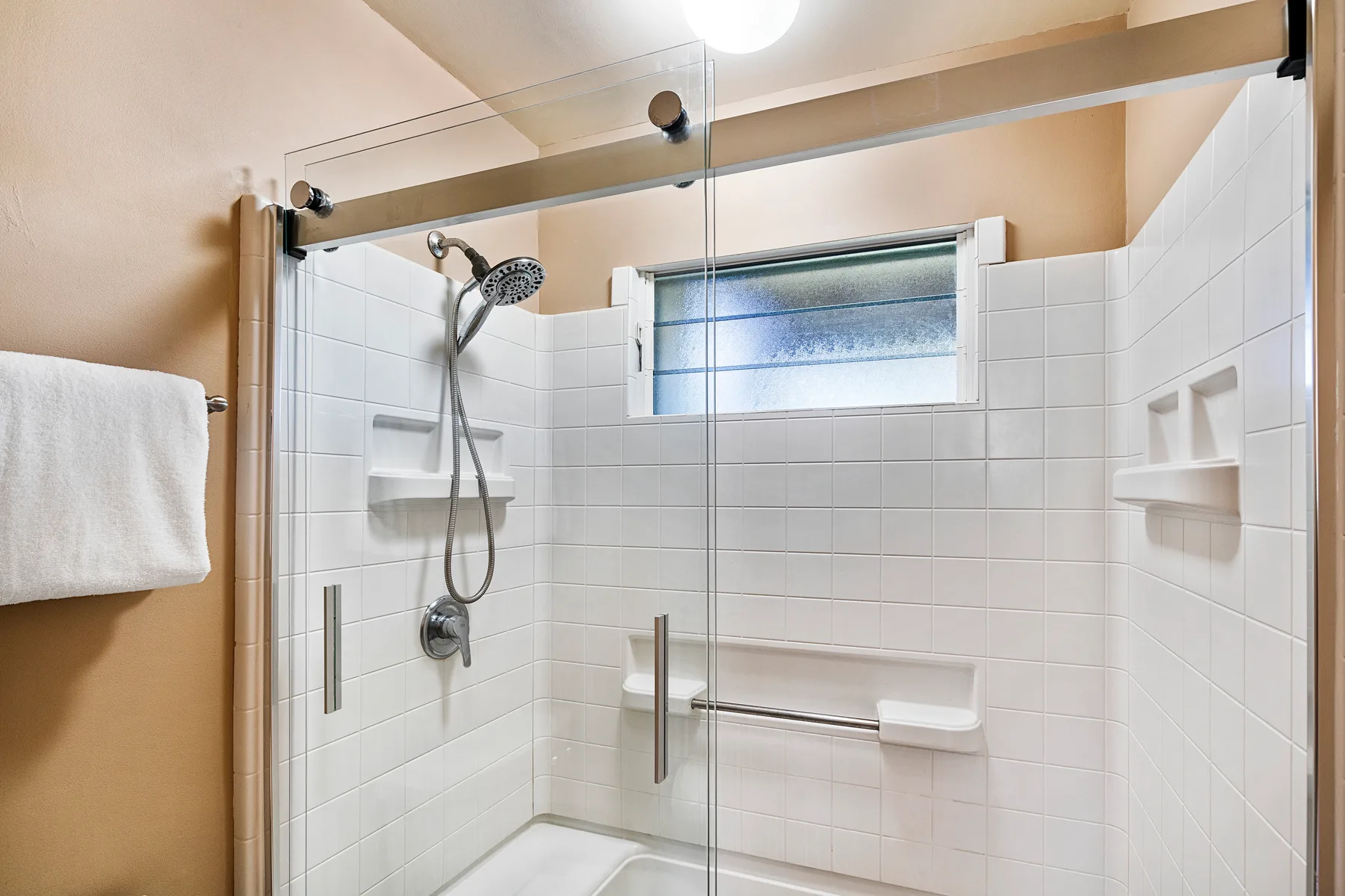 Un baño alicatado con ducha de vidrio, ducha de mano y una ventana pequeña.