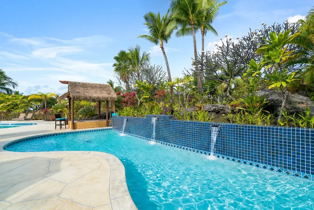 Una piscina exterior curva junto a un muro de azulejos y vegetación tropical.